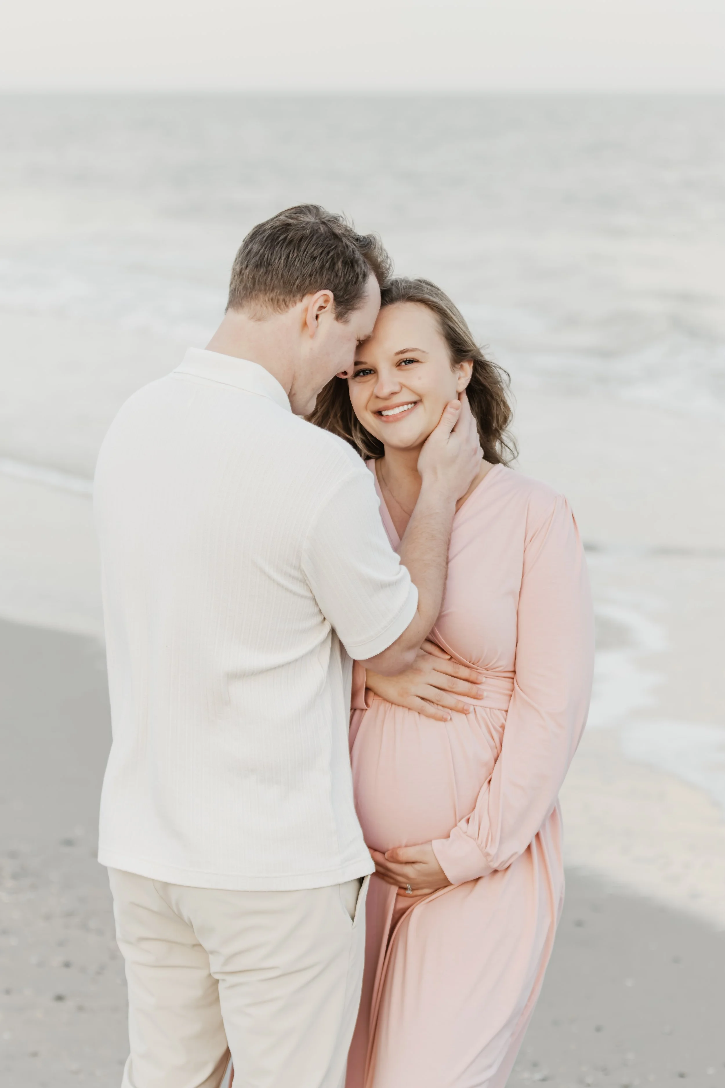 A smiling pregnant woman in a pink dress stands on the beach, with a man gently touching her face and neck. The beach has sand and ocean waves in the background.