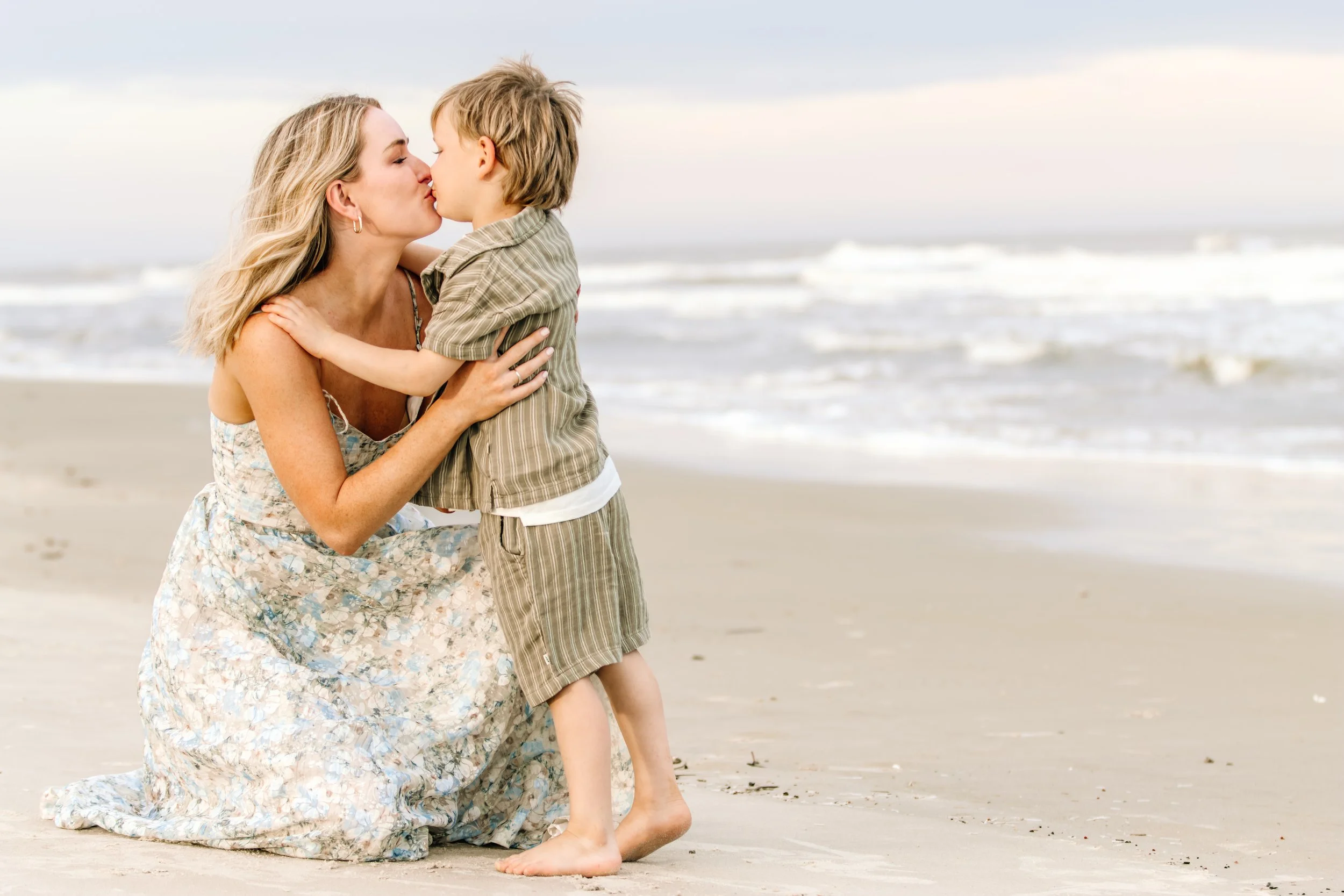 A woman kneeling on the beach, kissing a young boy, with ocean waves in the background on a cloudy day.