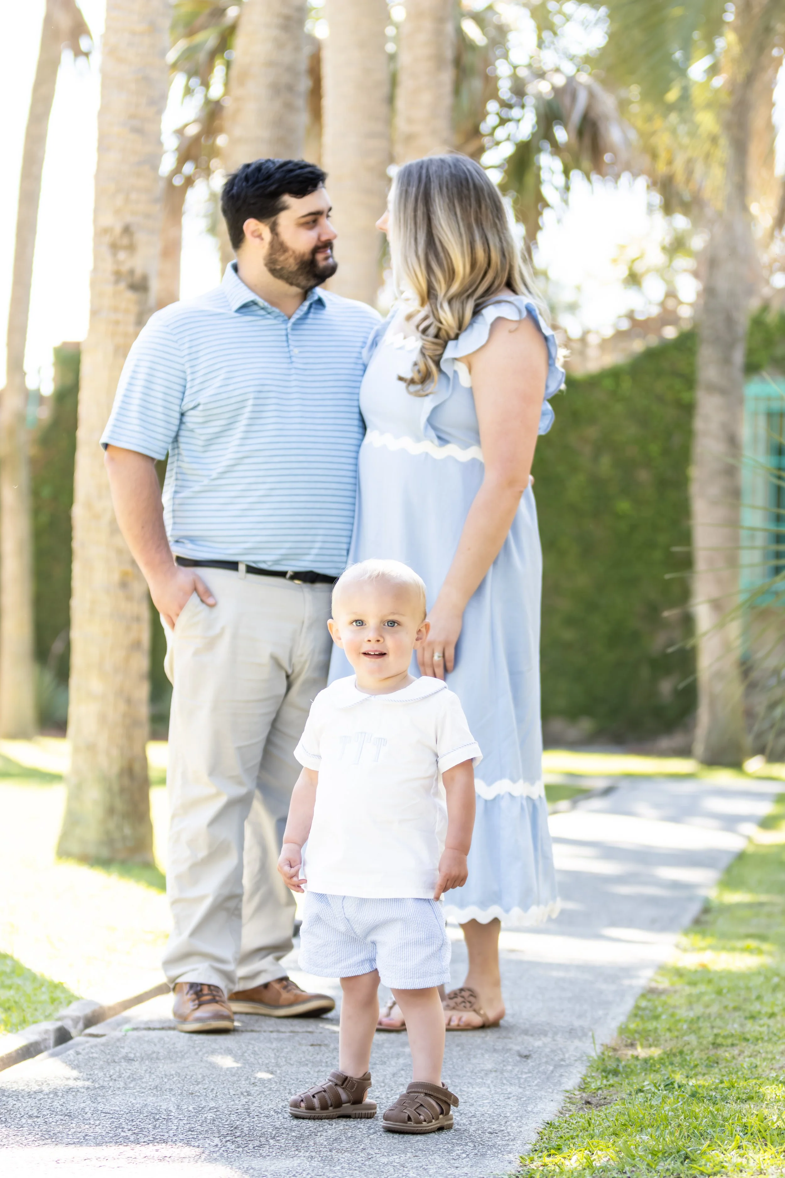 A family of three standing outdoors on a sidewalk with trees in the background. A pregnant woman, a man, and a young boy with blonde hair and light-colored clothing.