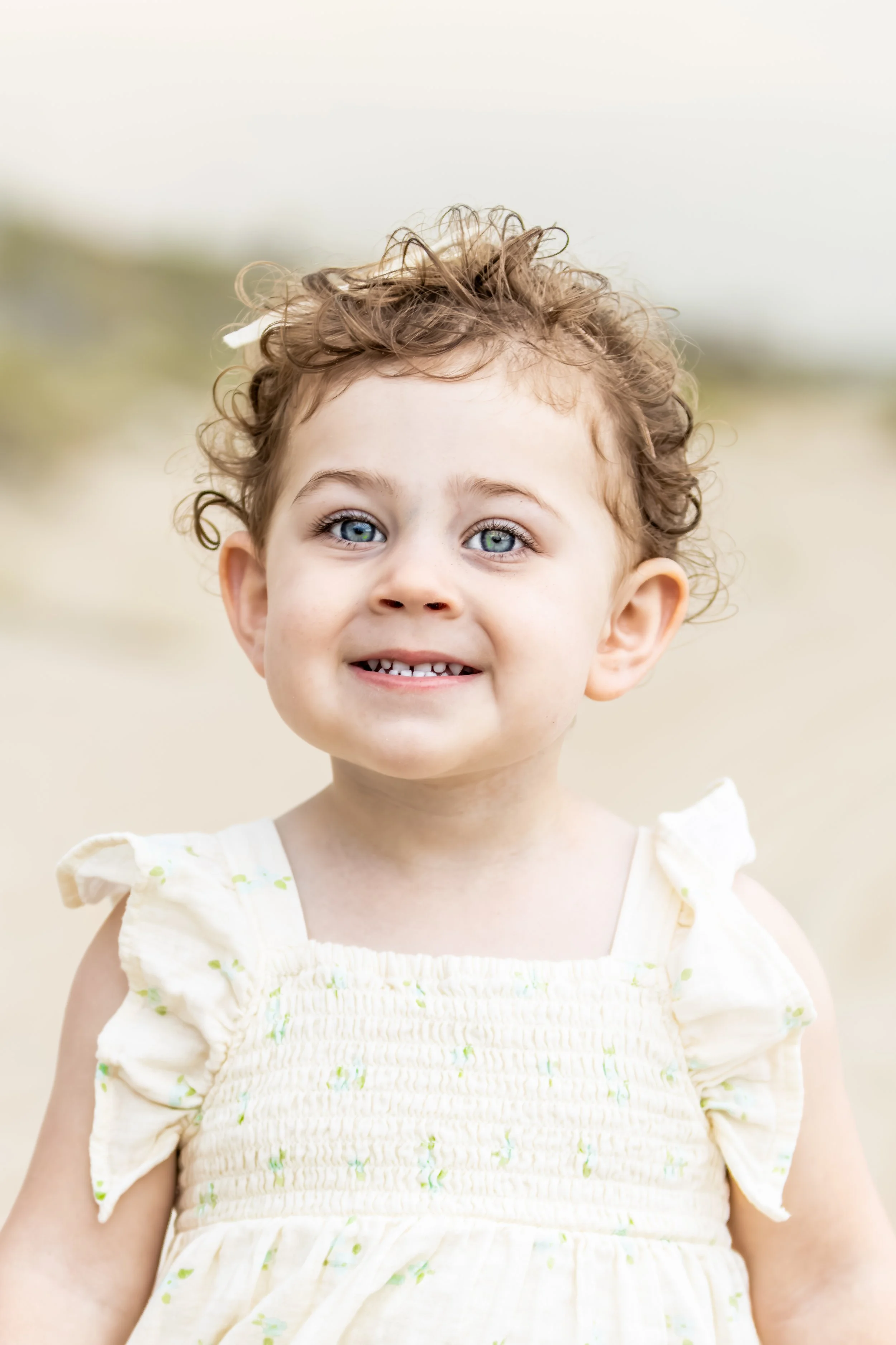 Young girl with curly brown hair and blue eyes smiling outdoors, wearing a light-colored dress with ruffled sleeves.