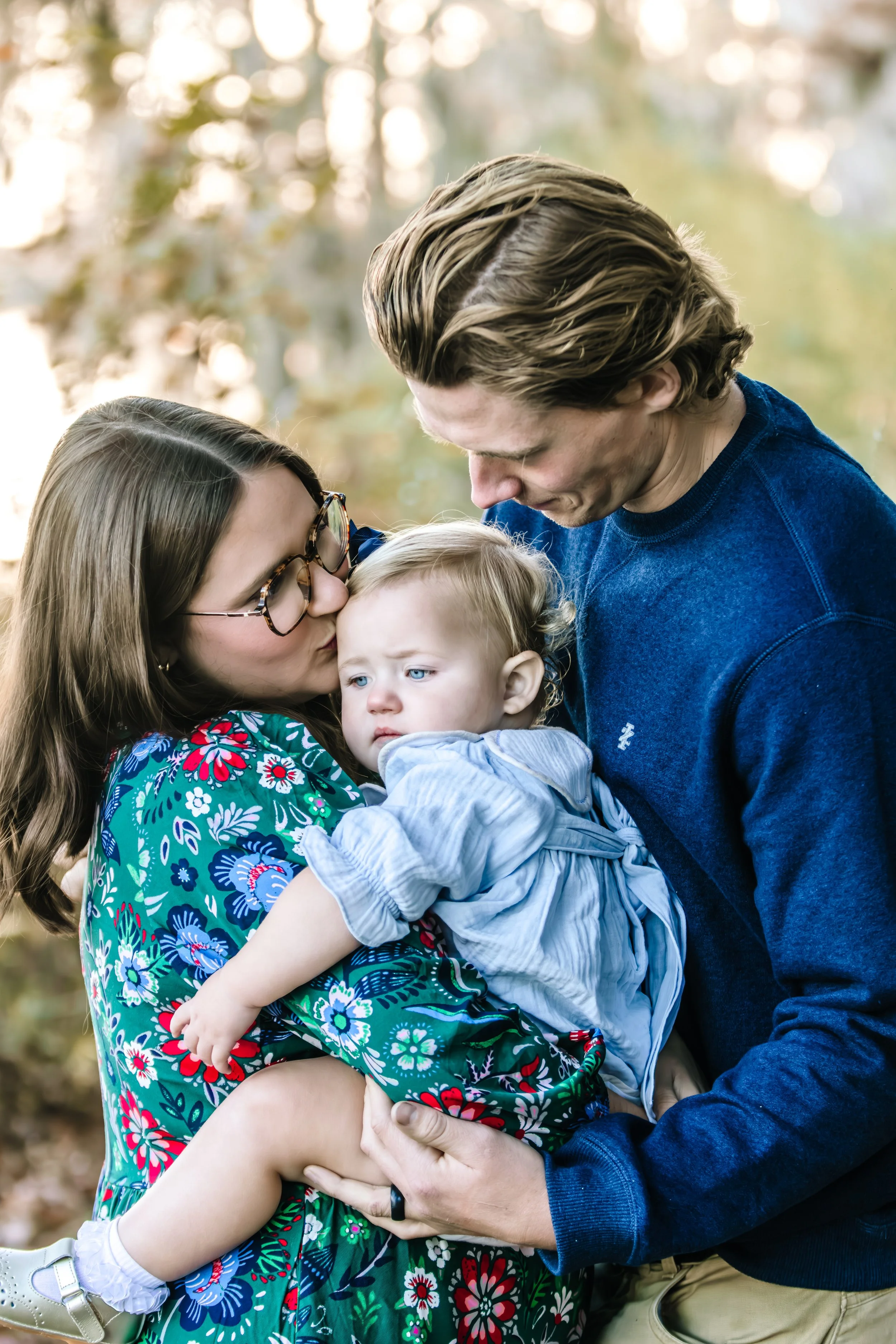 A woman holding a young child, with a man leaning over kissing the child's forehead in an outdoor setting with trees and sunlight in the background.