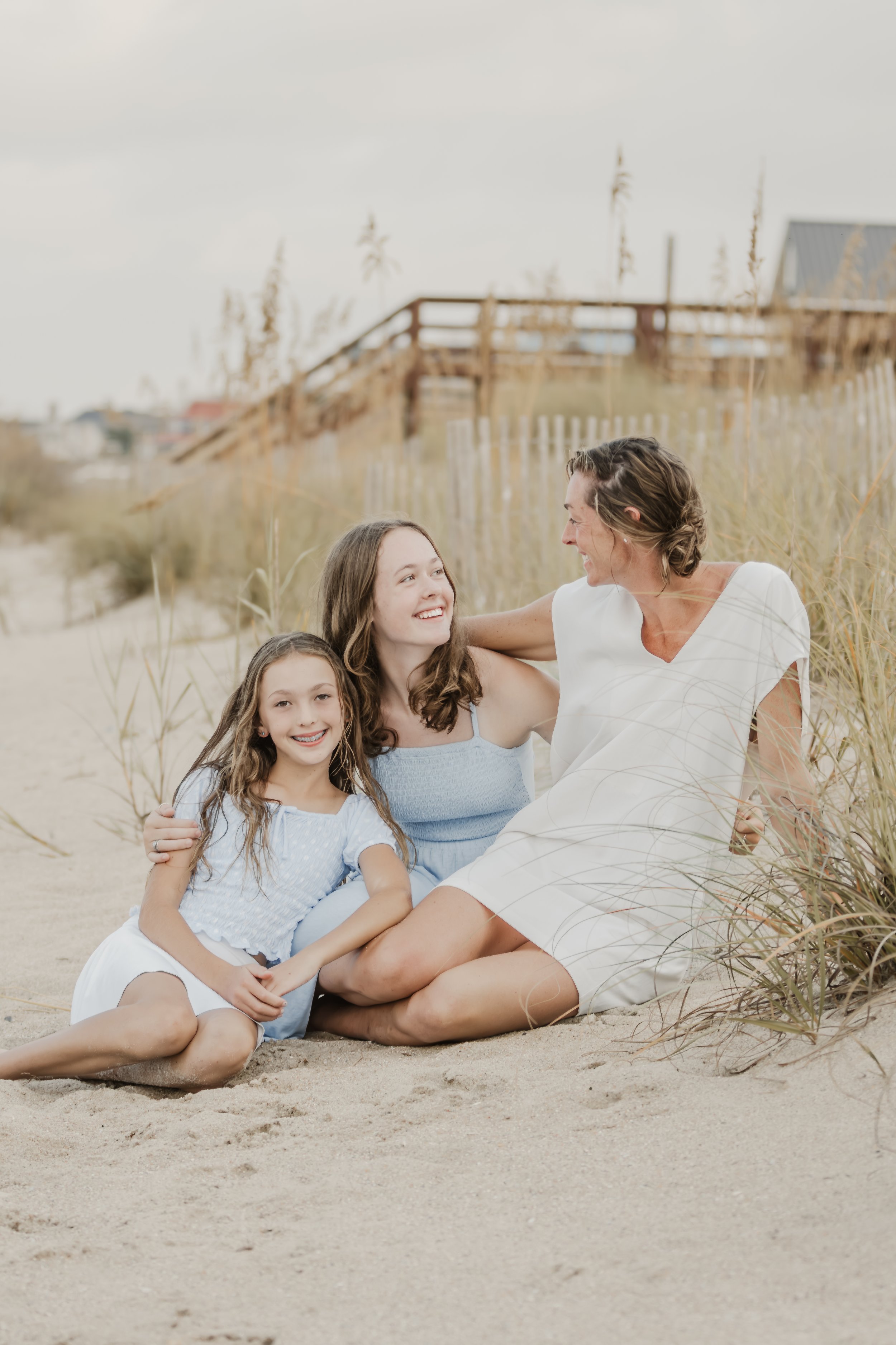 A woman and two girls sitting on the sand at the beach, smiling and looking at each other.