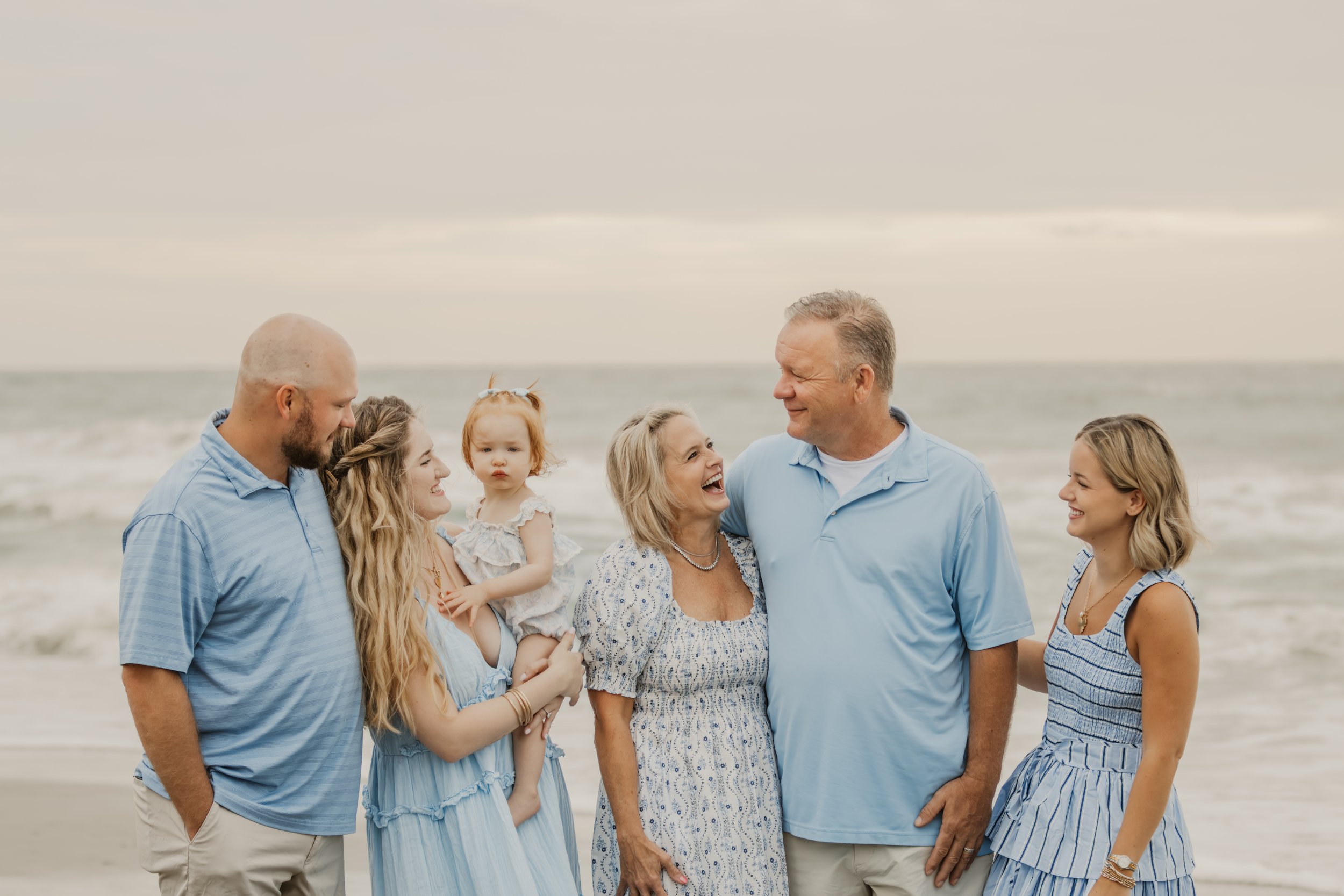 A family of six people standing on a beach, smiling and looking at each other. An older woman and man are in the center, the woman laughing. Three women and one man, along with a young girl, are around them, with the young girl sitting on one woman's