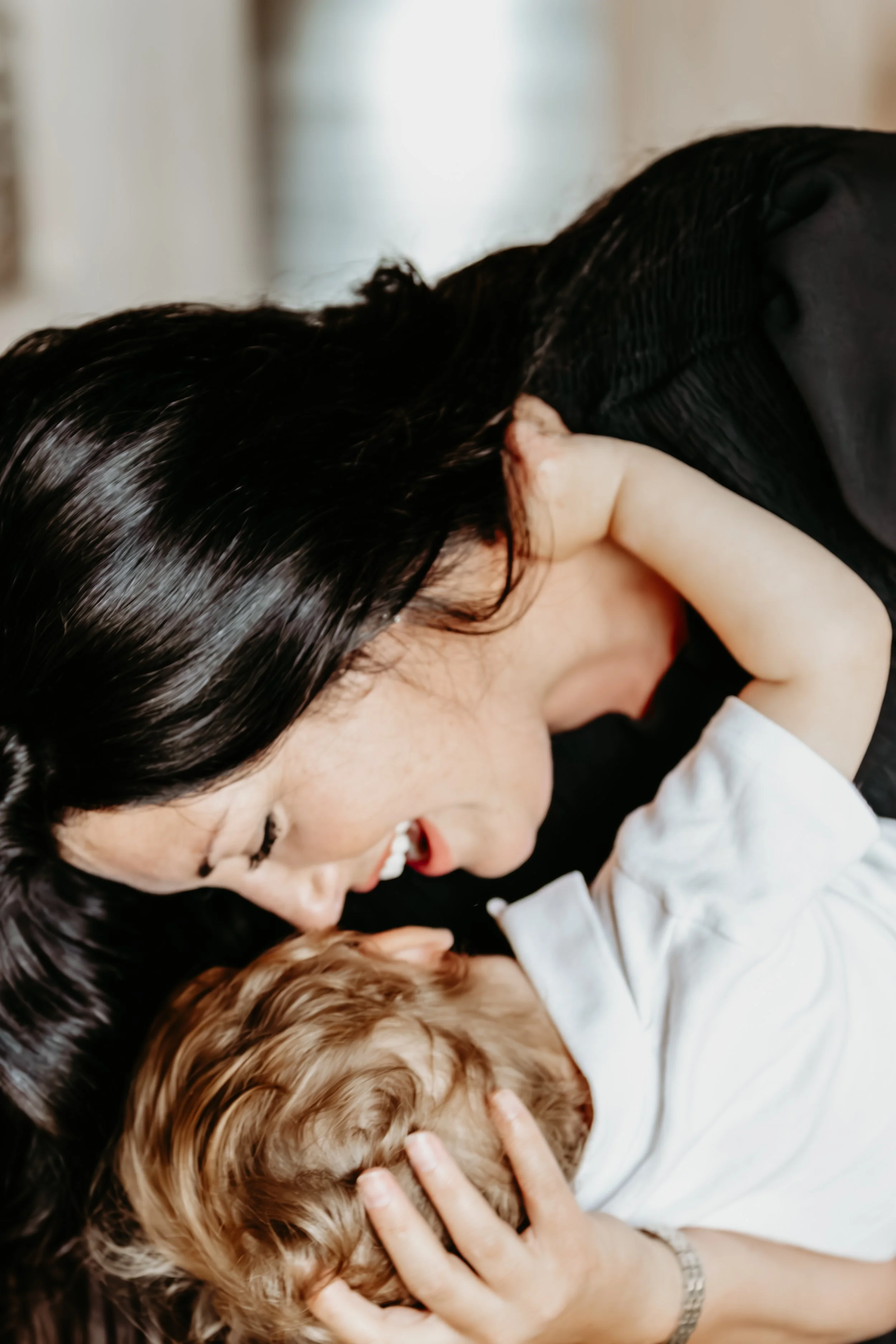 A woman with long dark hair is lying close to a child with curly hair, both of their faces touching affectionately.