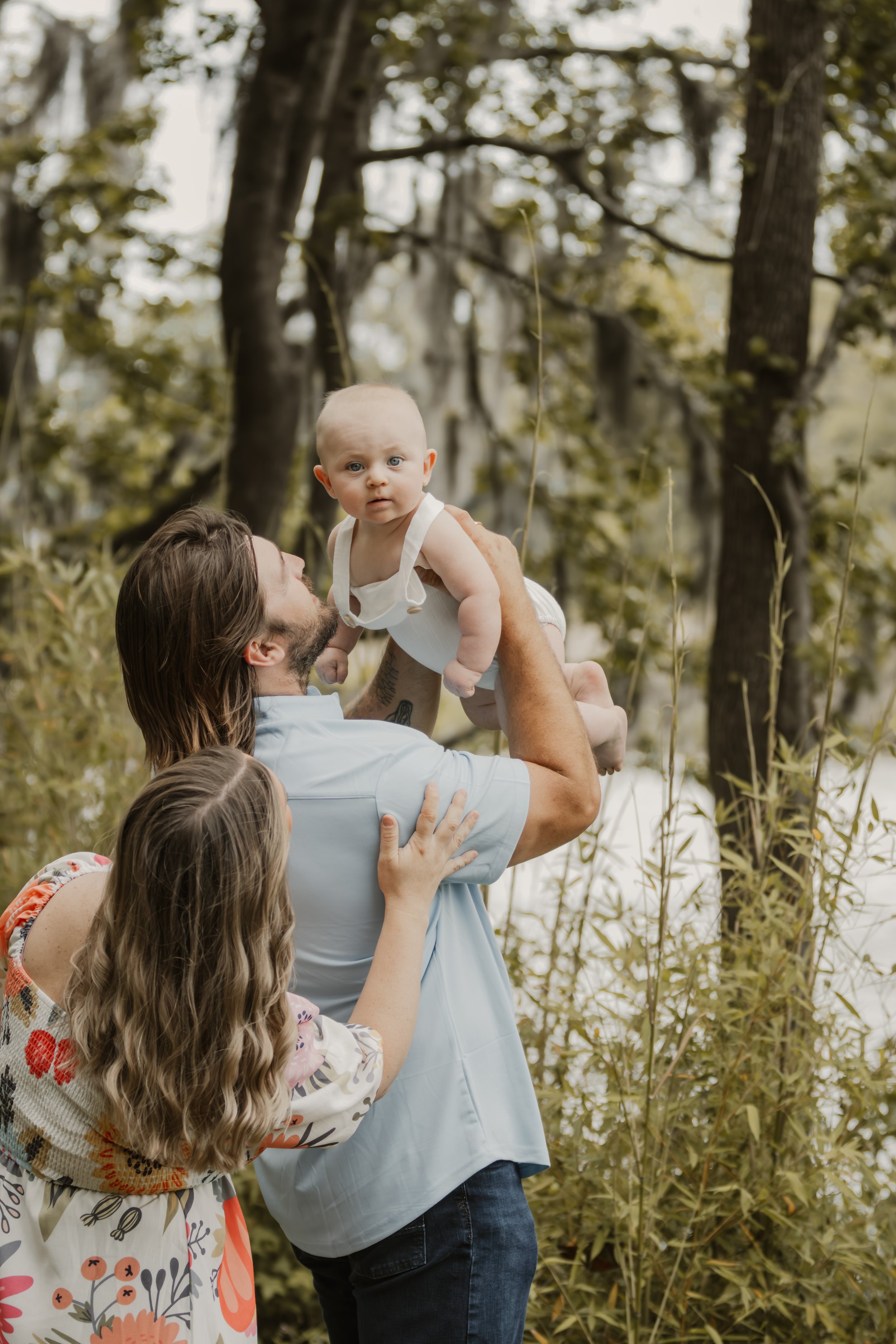 A man lifting a baby in the air while a woman and girl watch, outdoors near a lake and trees.