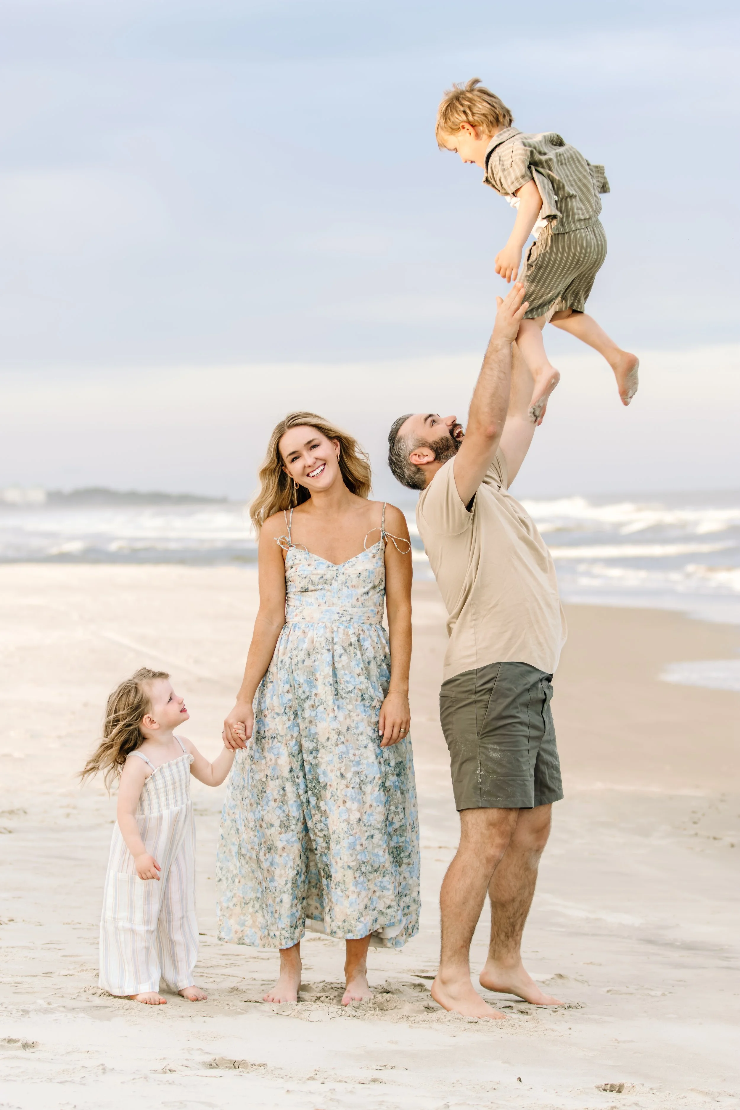 A joyful family on the beach, with a father lifting his young son into the air while a mother and daughter watch happily.