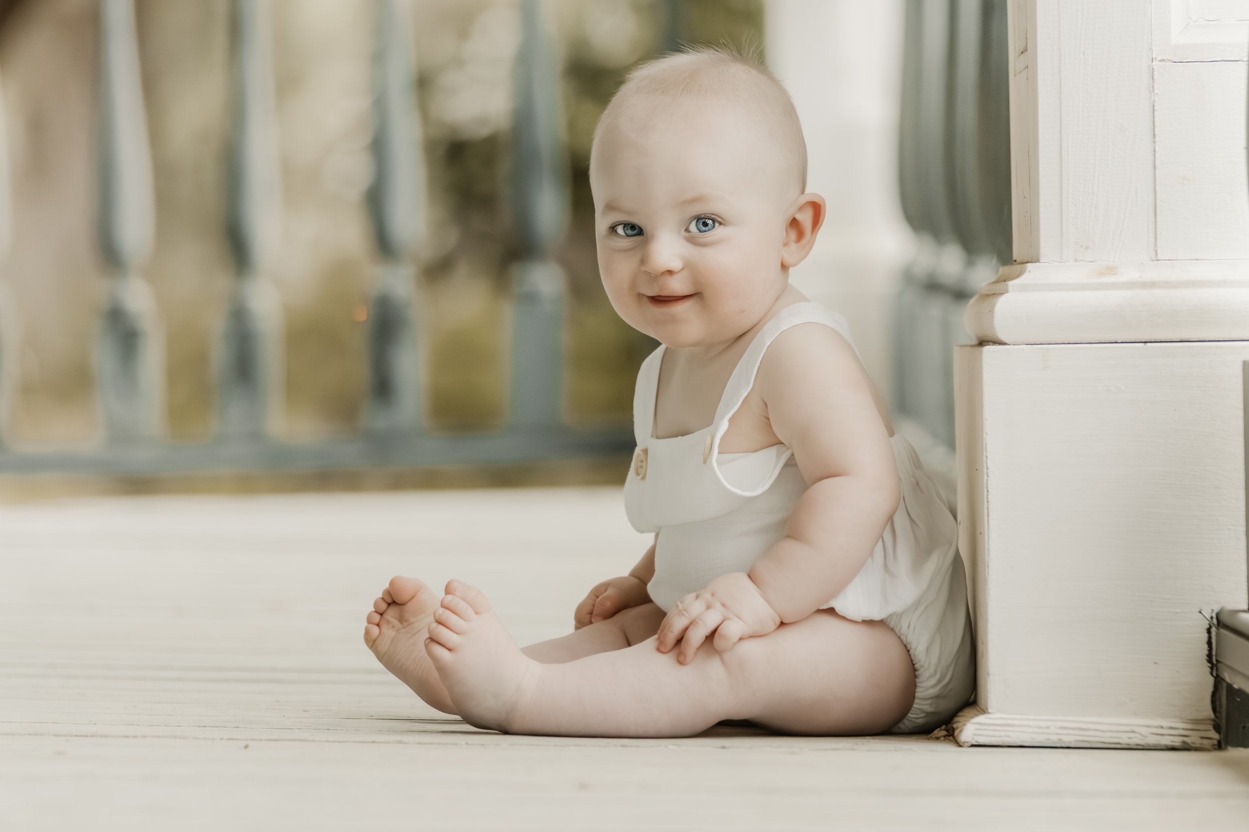 A smiling baby girl with blue eyes sitting on a wooden porch, wearing a white sleeveless dress, with a ruffled skirt and sitting near a white wooden structure.