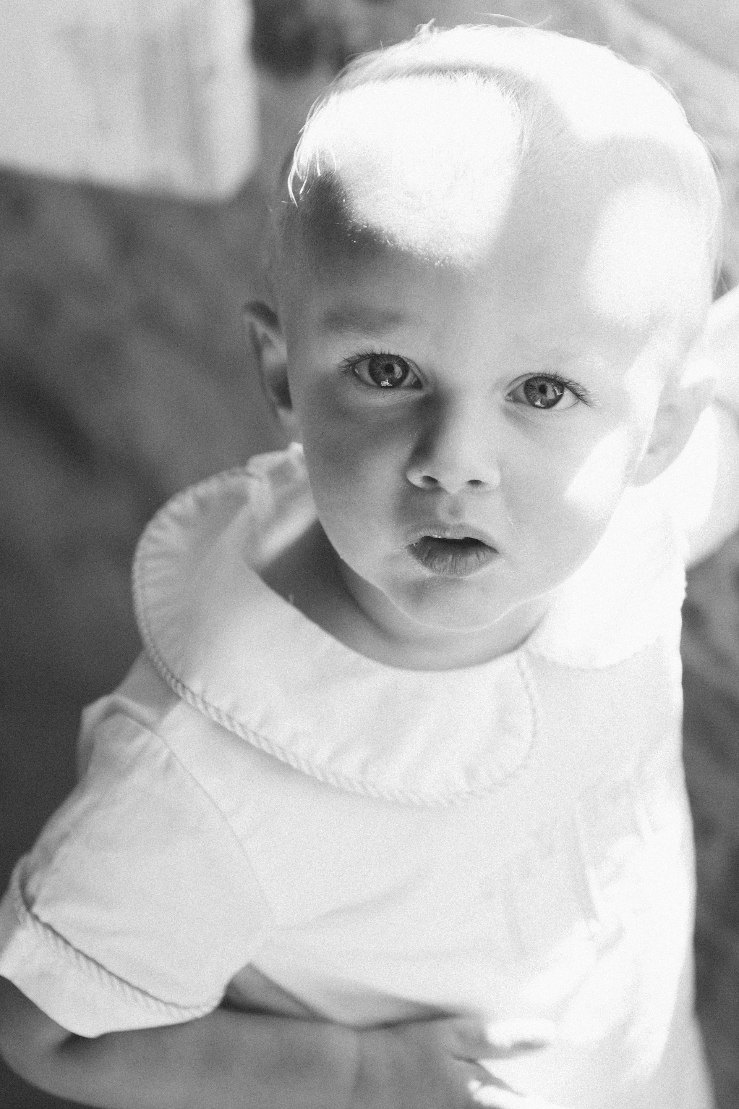 A young boy with light-colored hair and expressive eyes, looking up at the camera, wearing a light-colored shirt with a rounded collar, in black and white.