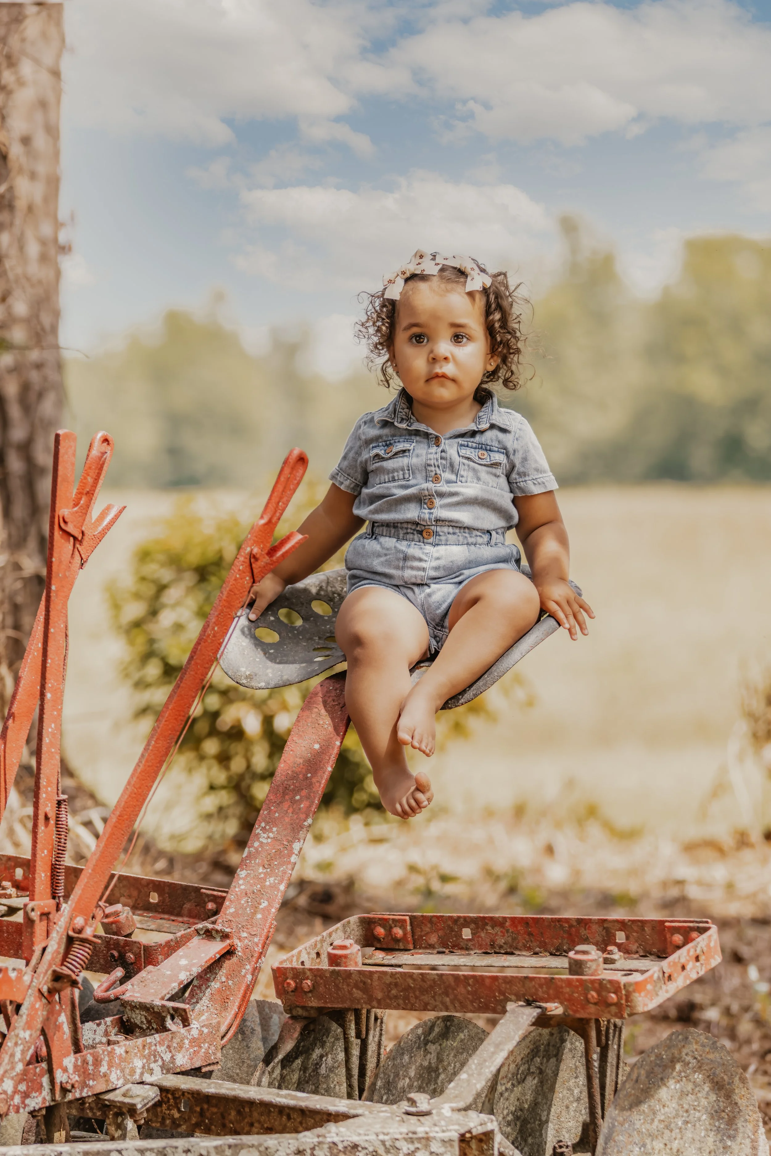 A young girl with curly hair, wearing a denim jumpsuit and a headband, sitting on an old, rusty farm implement outdoors in a rural setting with trees and a blue sky in the background.