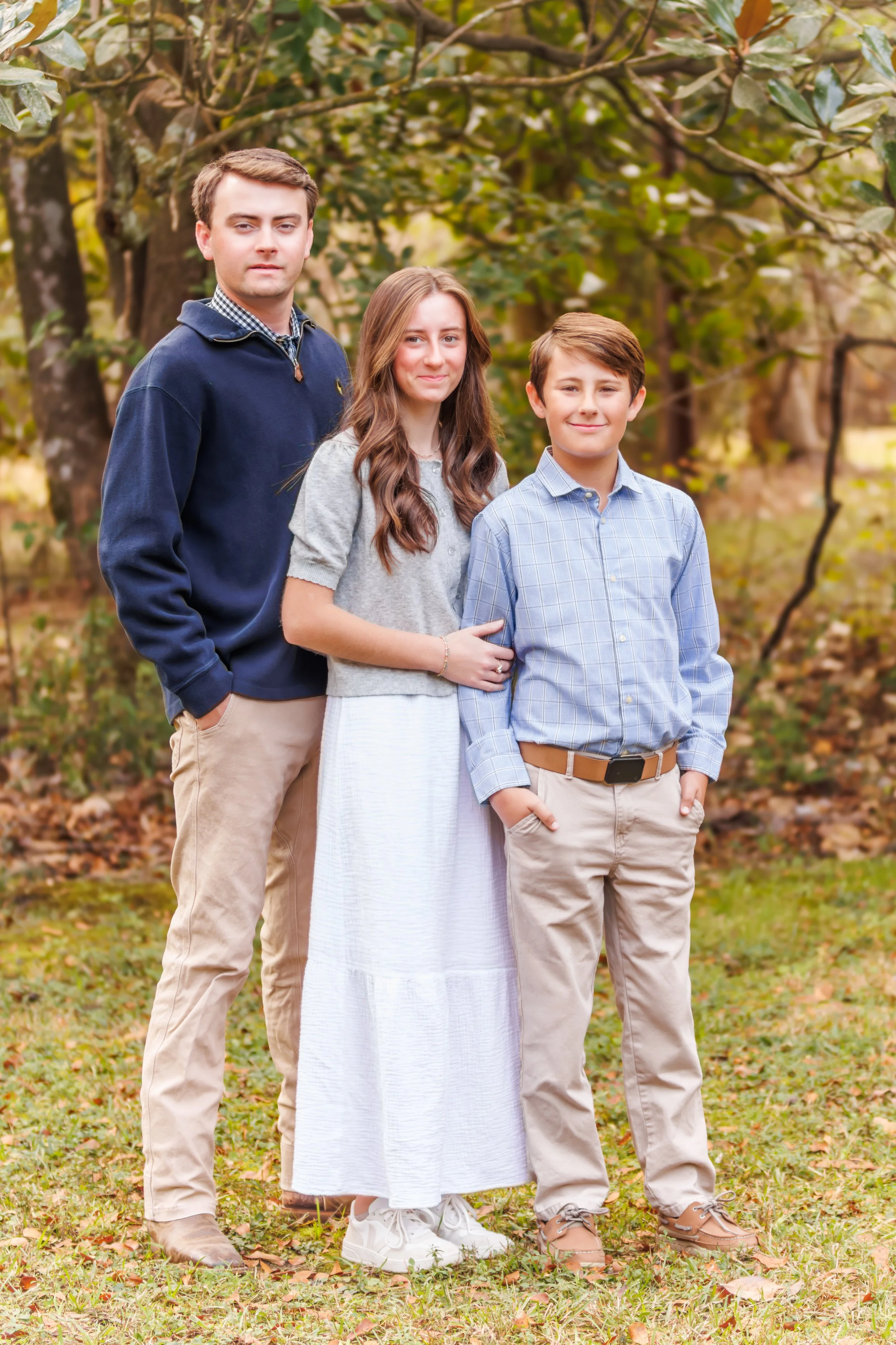 Three young people standing outdoors in a park with fallen leaves and trees in the background.