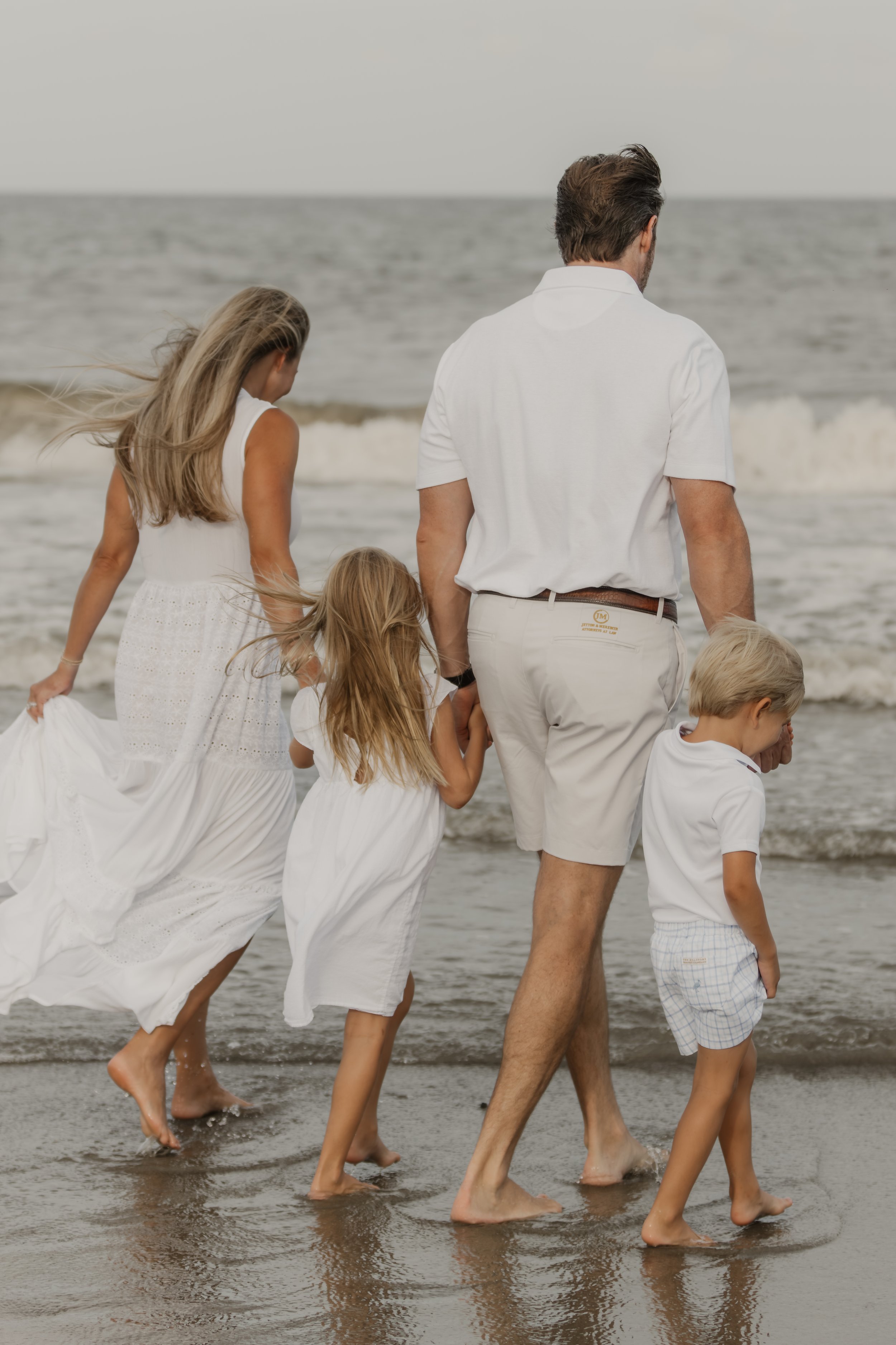 Family walking along the shoreline at the beach, dressed in white, with waves in the background.