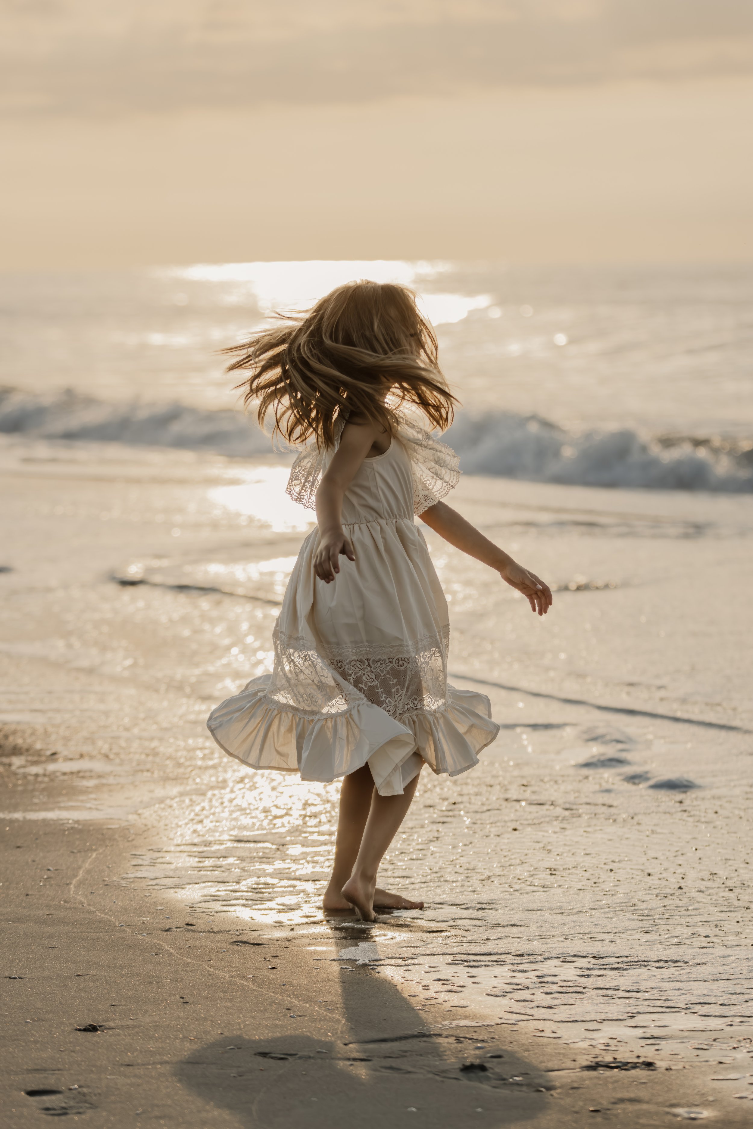 A young girl in a white dress walking along the shoreline at sunset with her hair blowing in the wind.