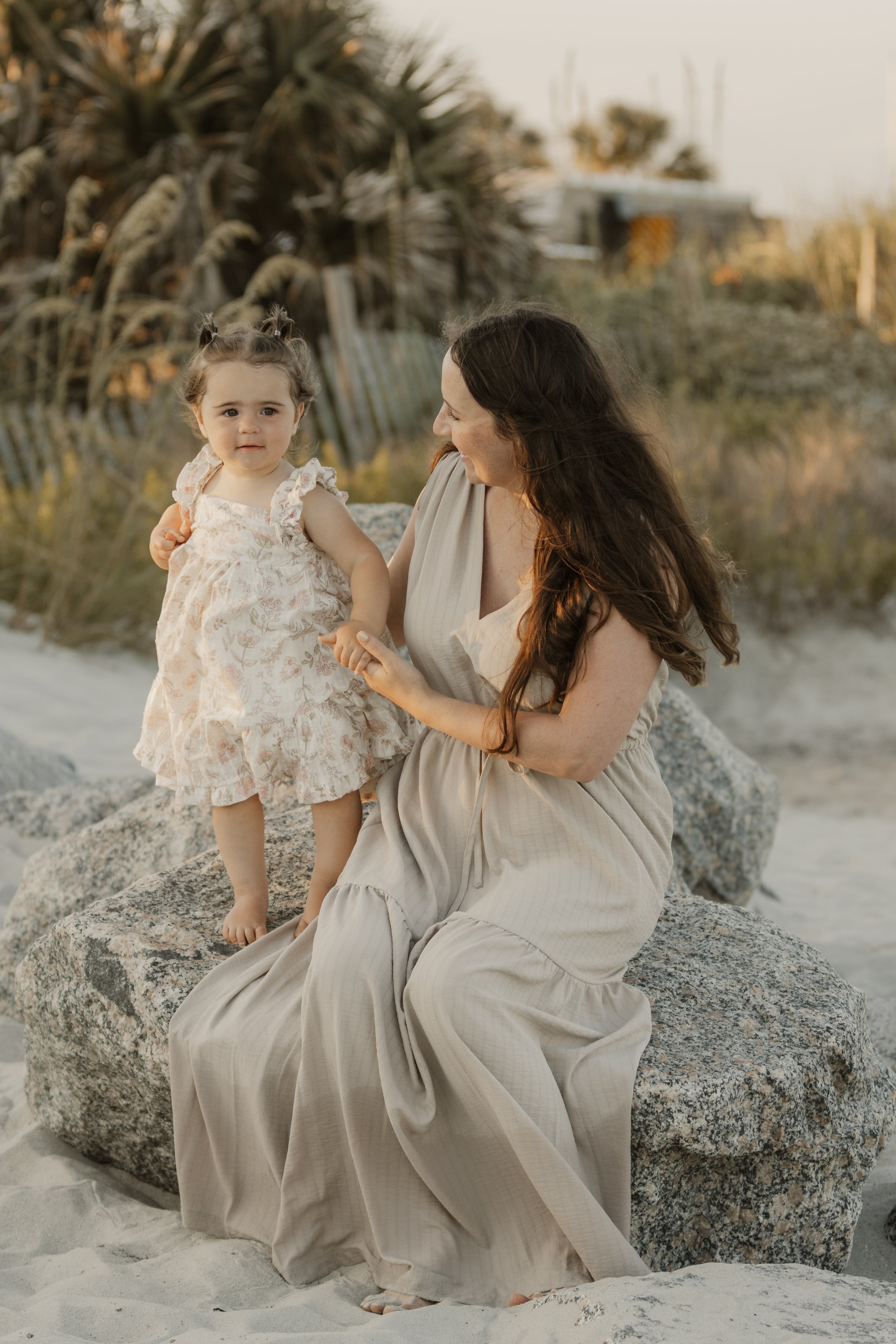 A woman and a toddler girl sitting on rocks at the beach during sunset, with sand, rocks, and beach vegetation in the background.
