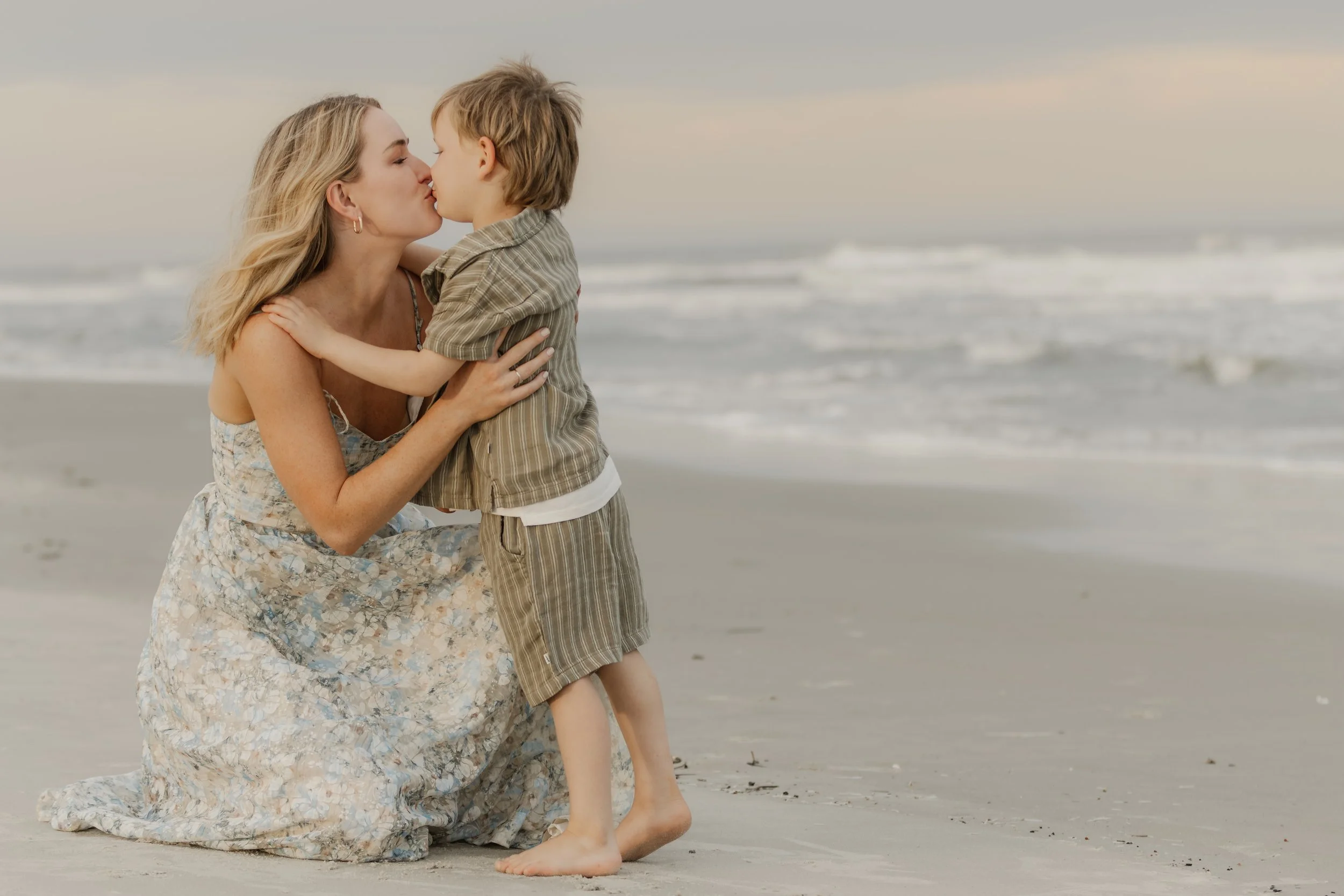 A woman and a young boy share a kiss on the beach, with waves in the background during sunset.