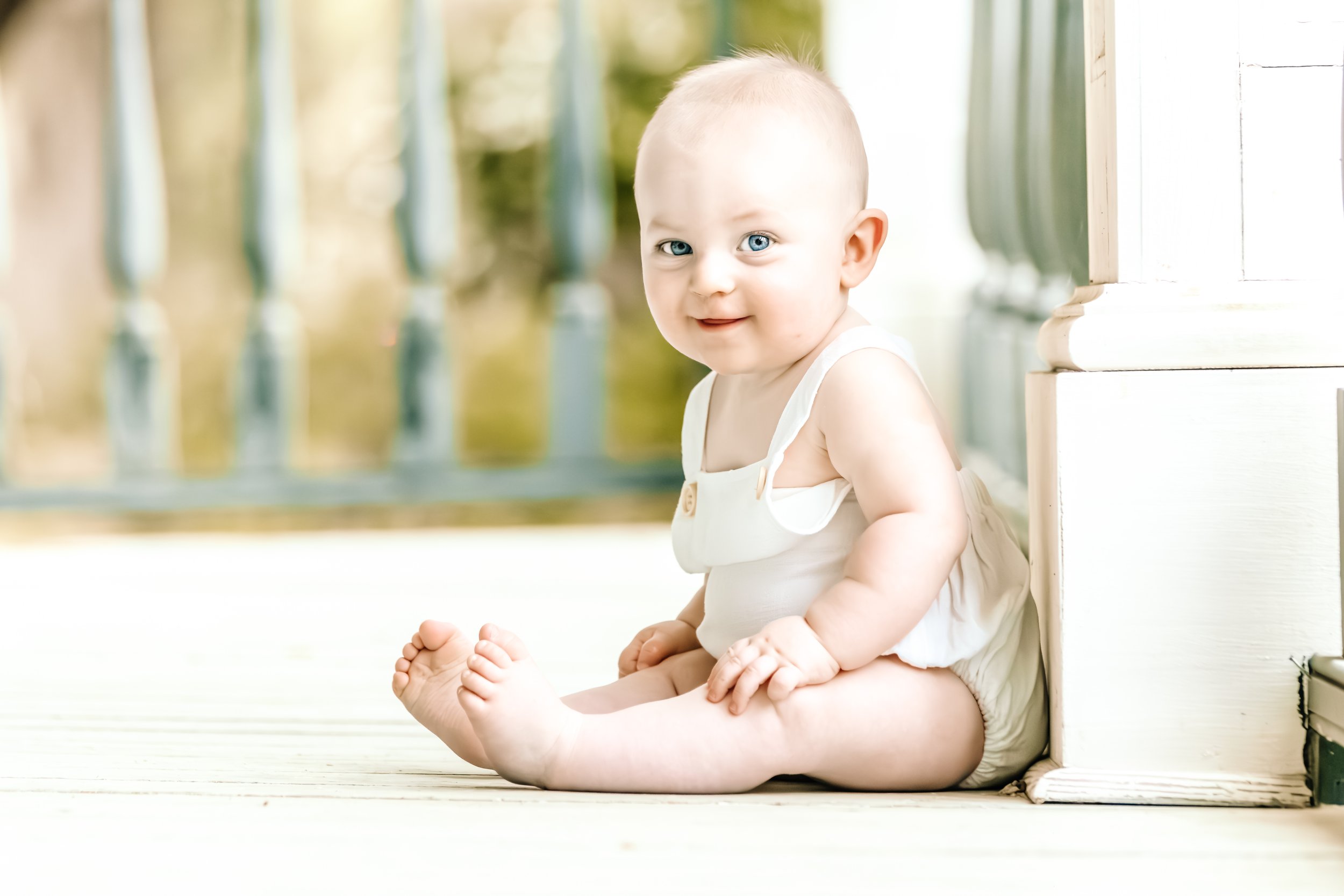 A smiling baby with blue eyes, sitting on a porch with a white wooden floor, wearing a white sleeveless outfit. The baby is sitting next to a white wall and looking at the camera.