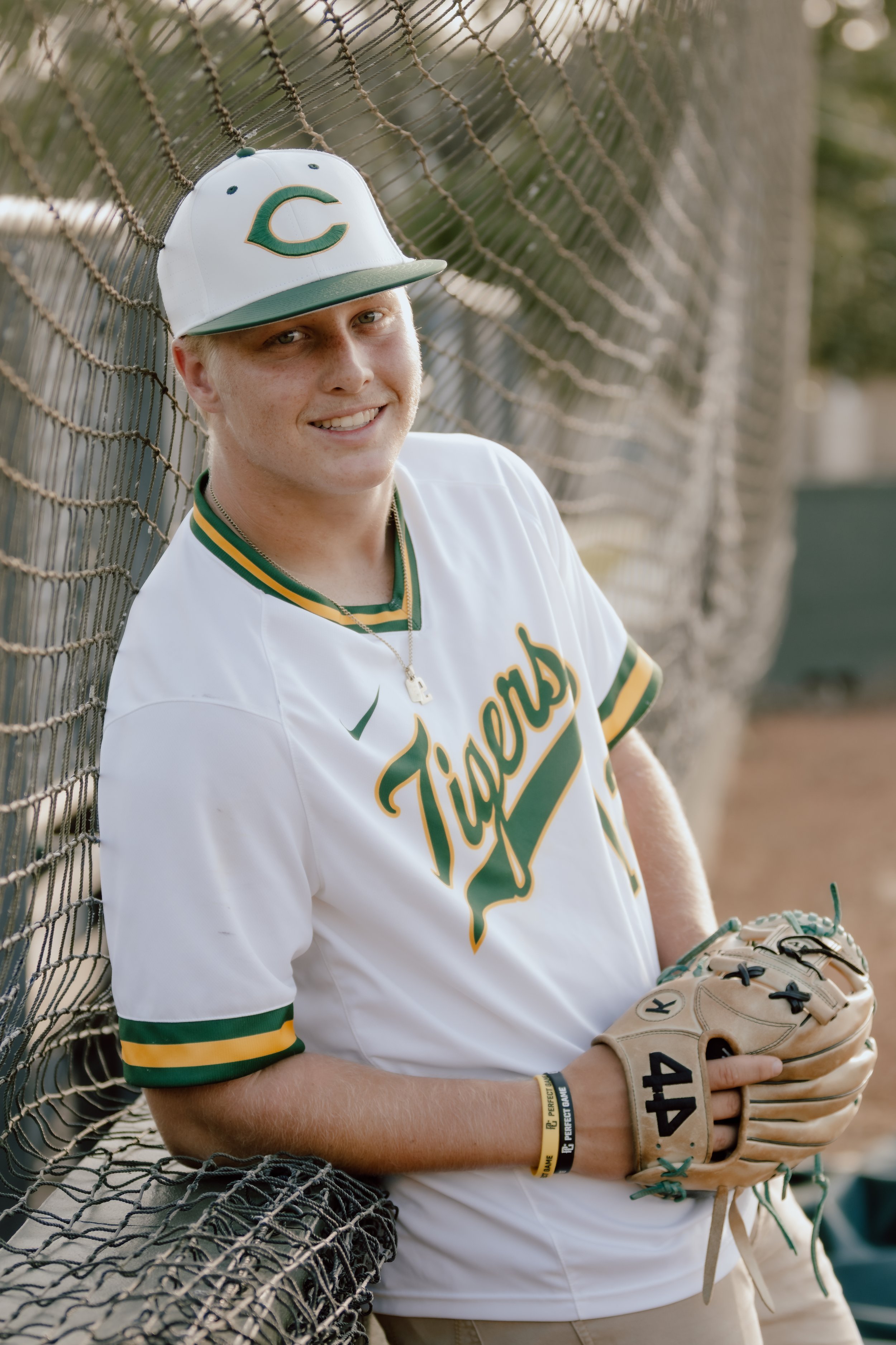 A young man in a white and green baseball uniform with 'Tigers' written on it, wearing a baseball cap, sitting on a bench against a chain-link fence, holding a baseball glove and smiling at the camera.