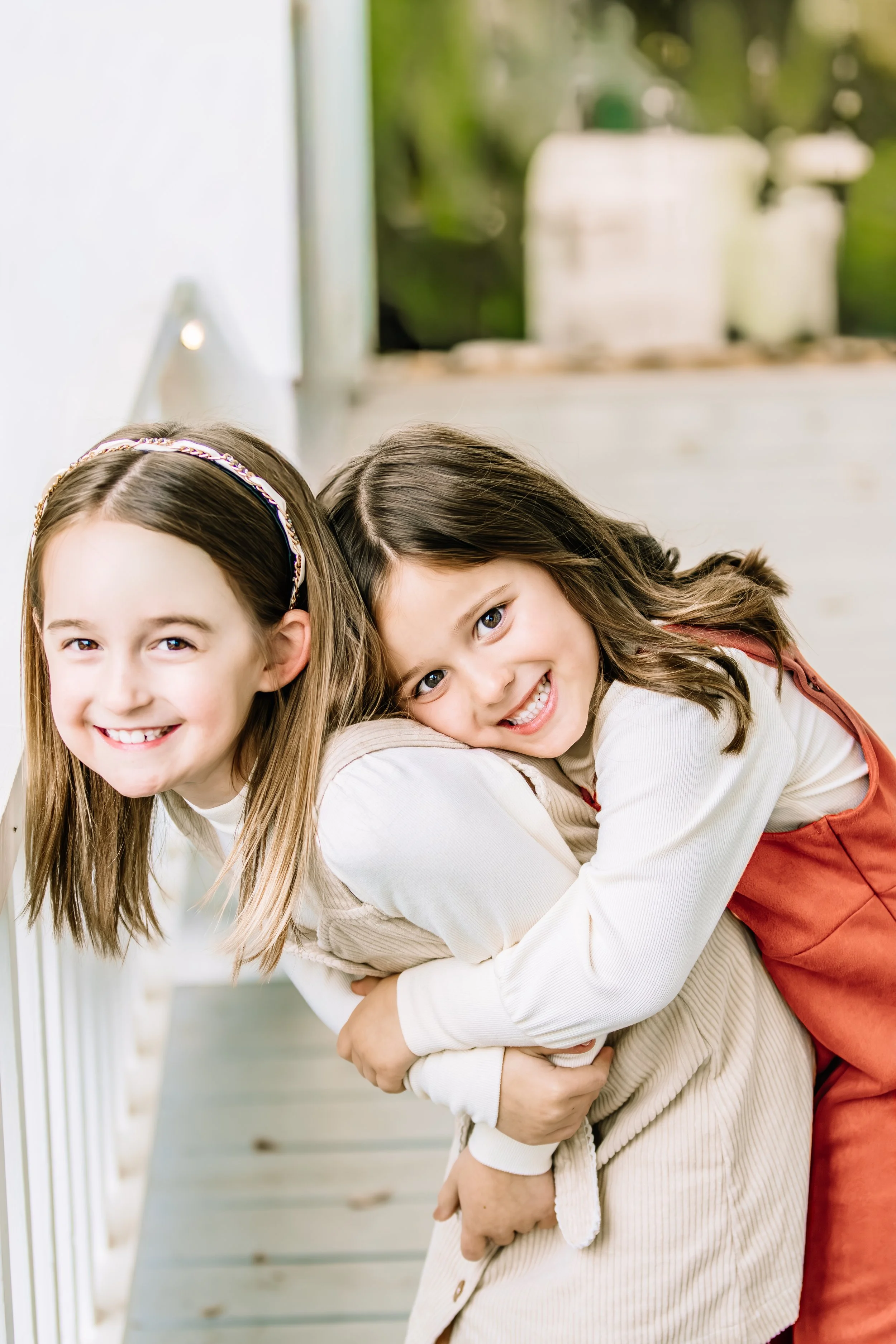 Two young girls smiling, hugging each other on a porch with white railings and a blurred background of greenery.
