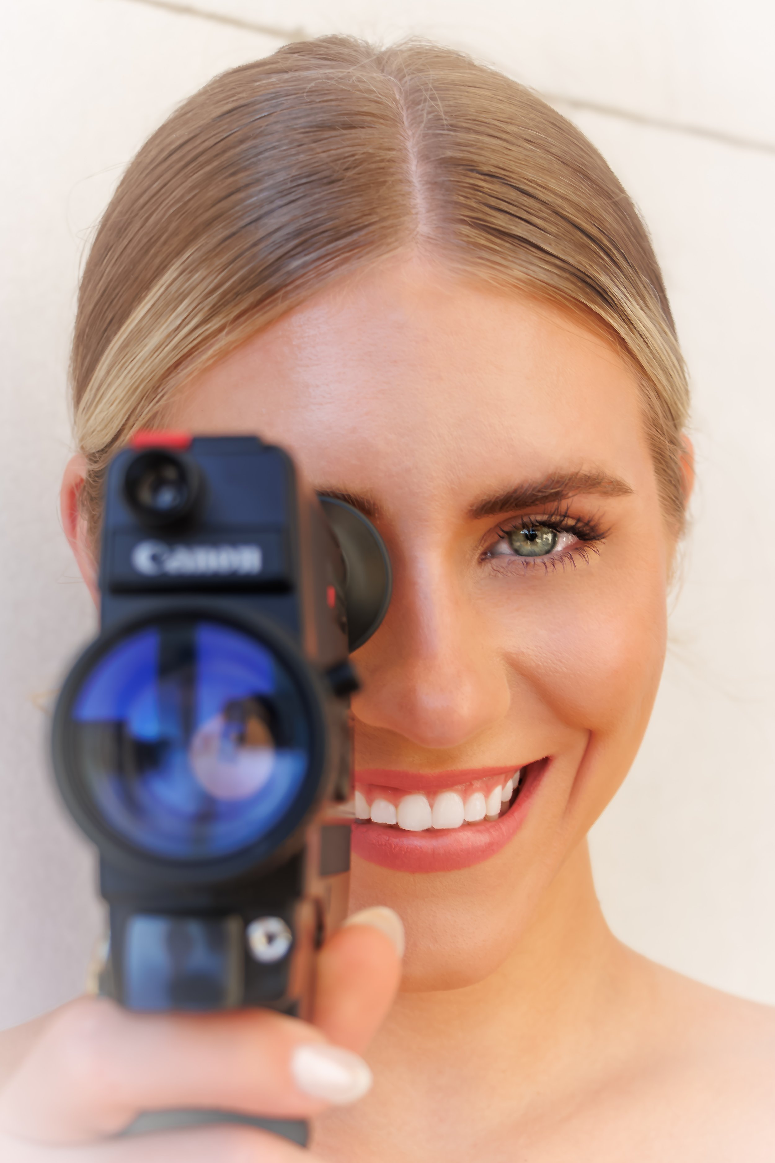A smiling woman with blonde hair and green eyes, looking through a camera's scope to the viewer.