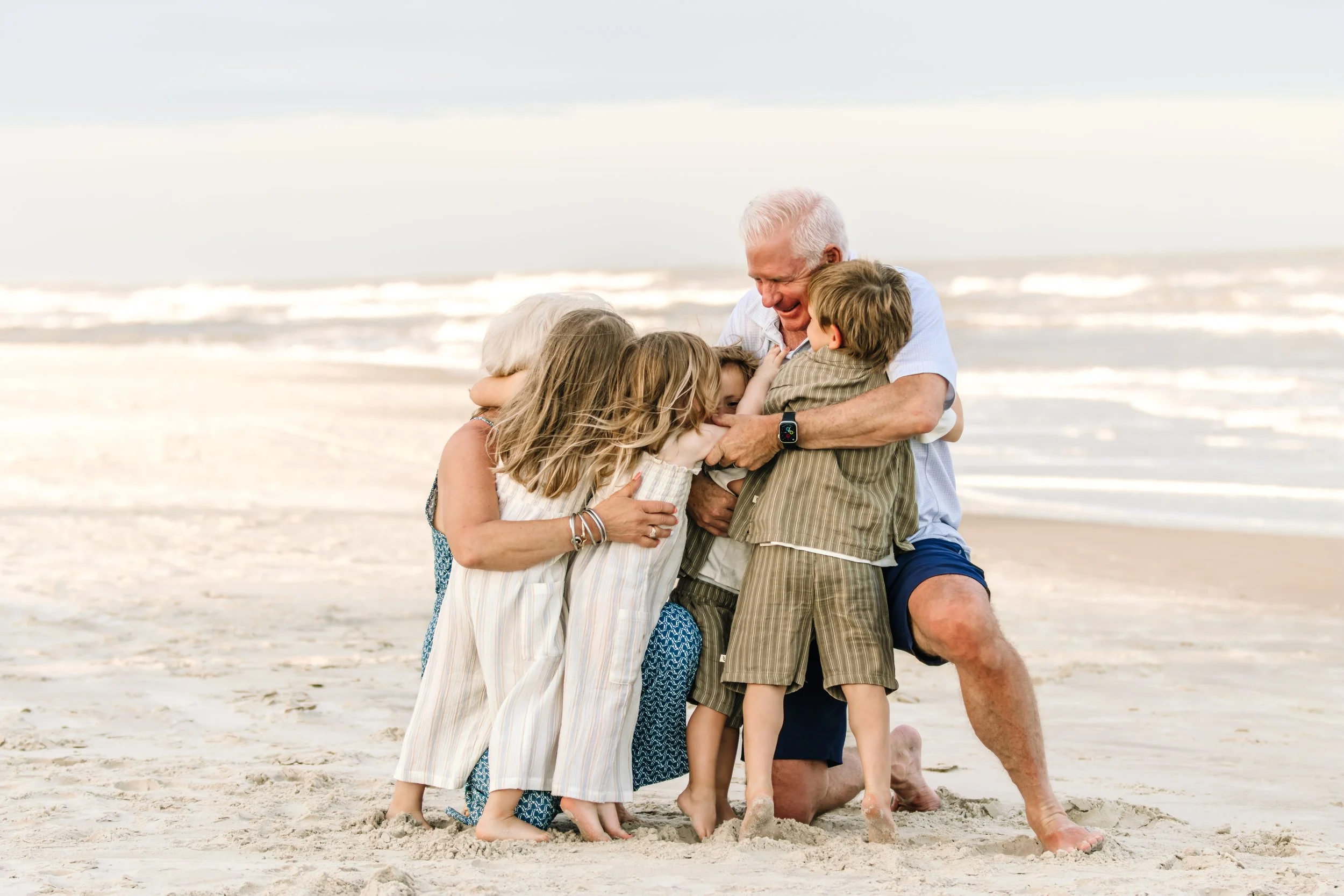 An elderly man and woman with four children hugging on a sandy beach near the ocean.