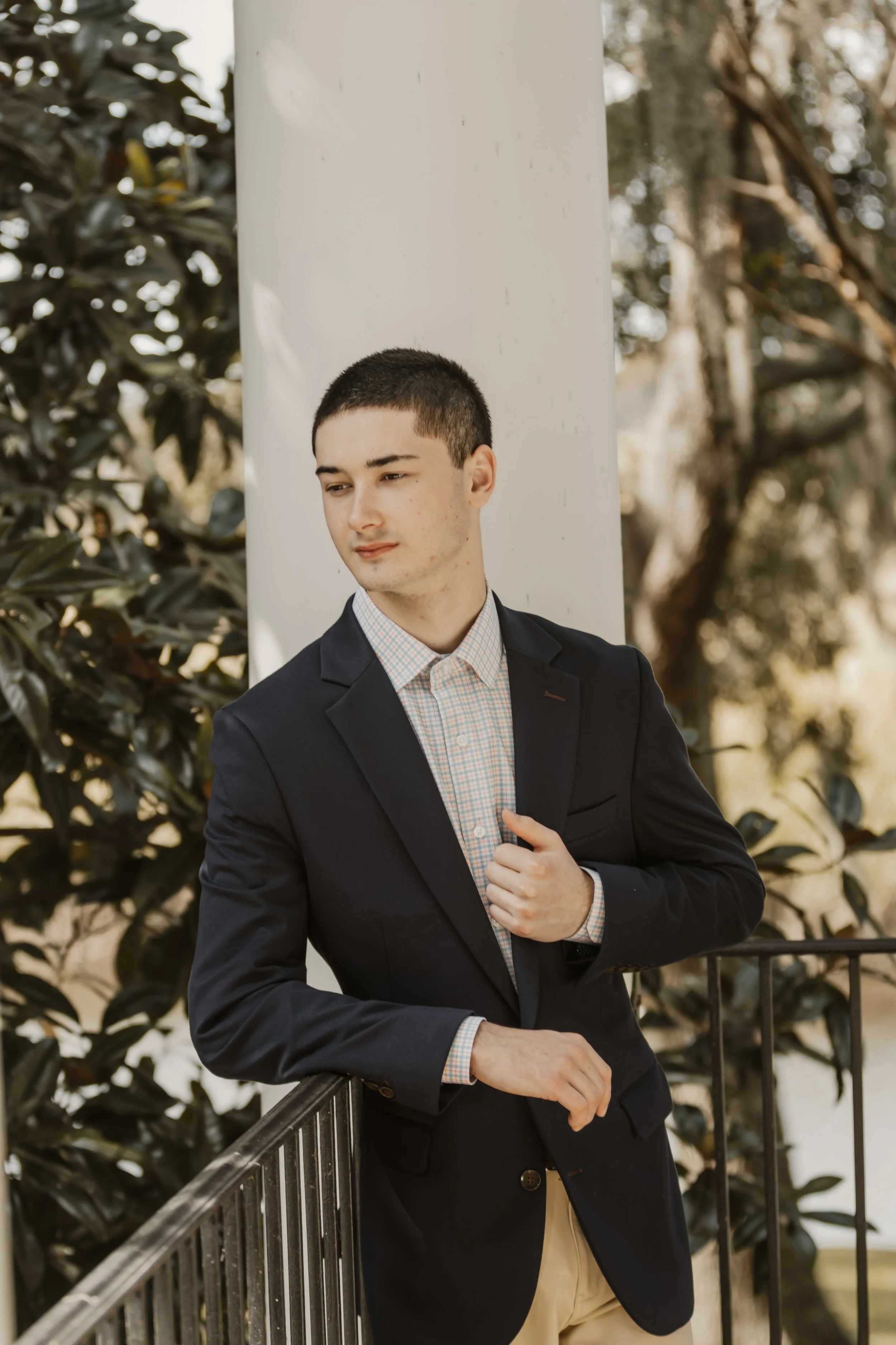 A young man in a dark blazer and checkered shirt leaning against a black railing outdoors with trees and foliage in the background.