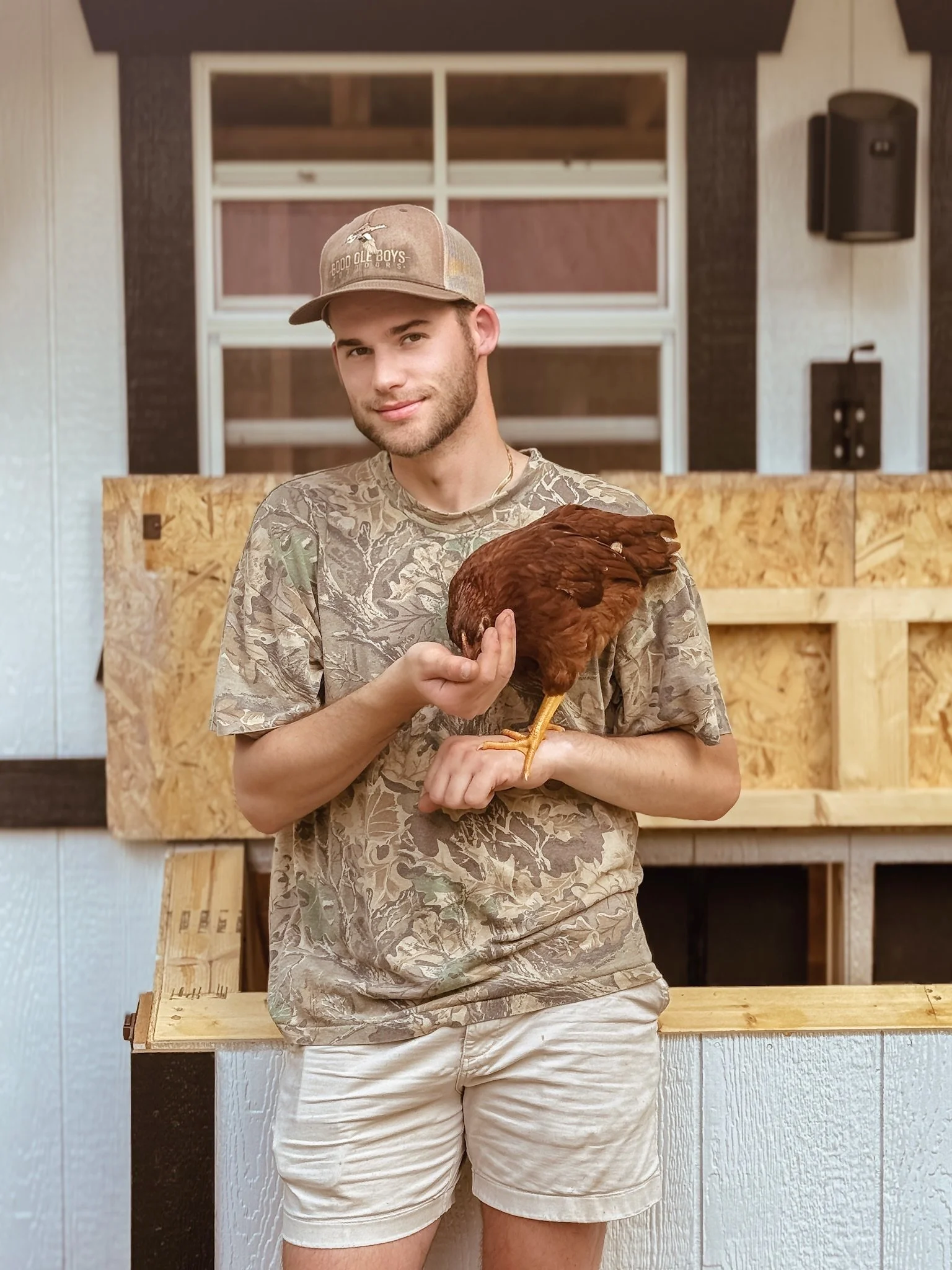 A young man wearing a desert camouflage t-shirt, beige shorts, and a tan baseball cap with text, standing indoors and holding a brown chicken.