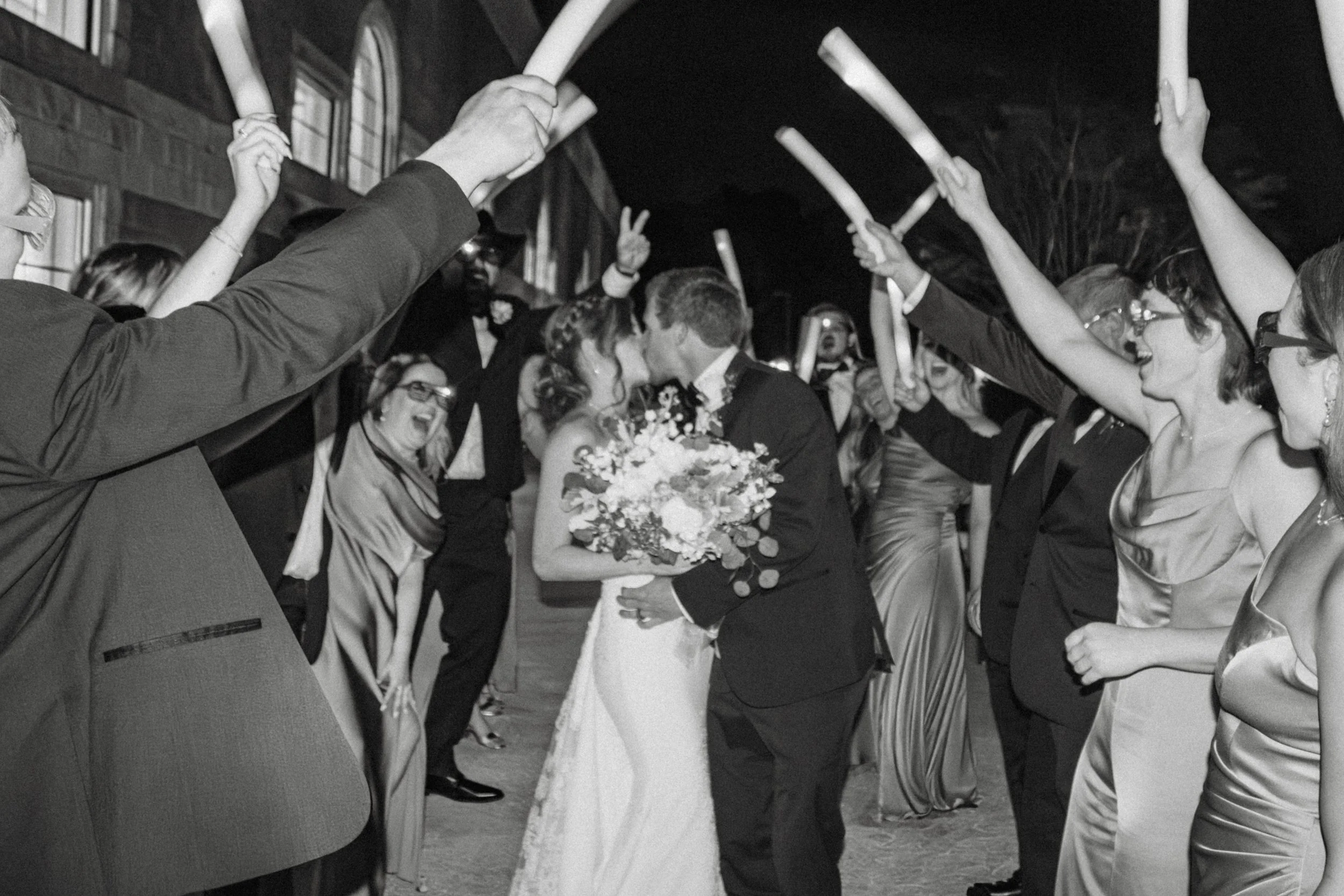 Black and white photo of a bride and groom sharing a kiss at their wedding reception, surrounded by guests raising glow sticks in celebration.