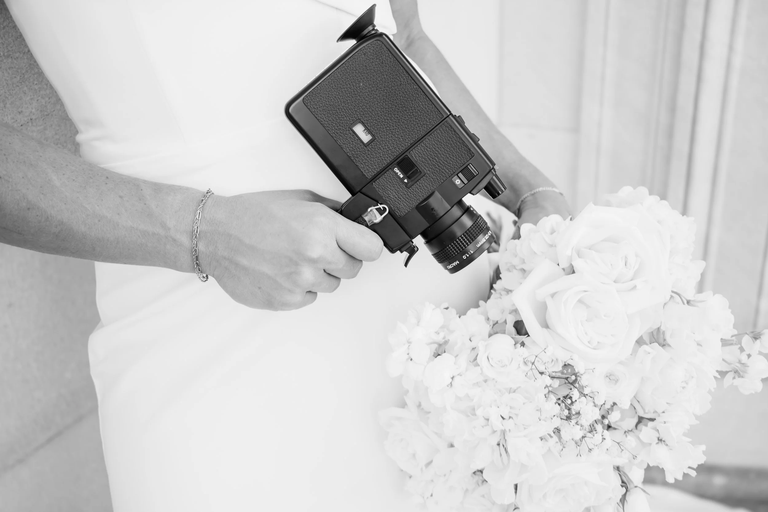 Person holding a vintage camera while holding a bouquet of white roses, wearing a white dress and a bracelet.