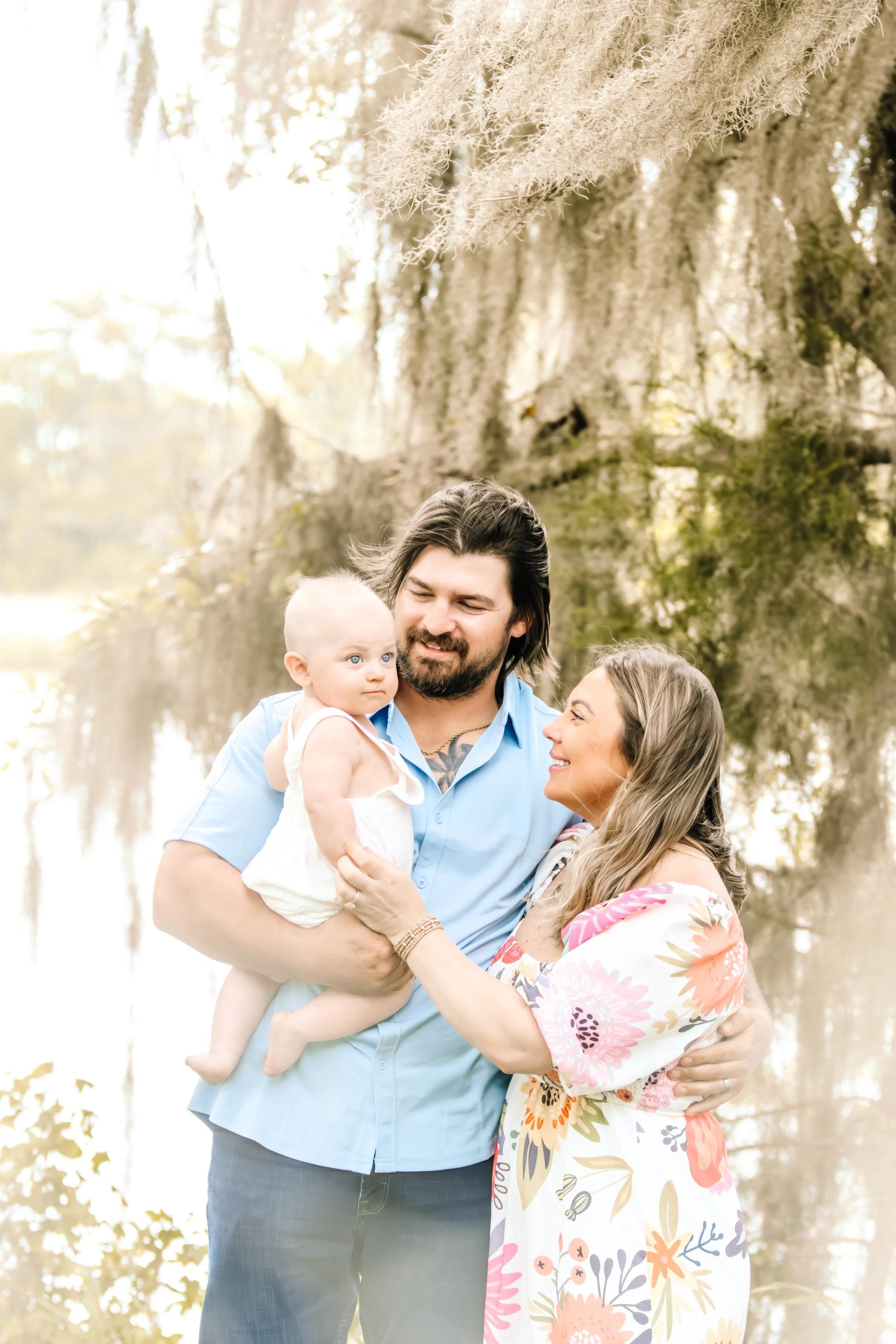A family of three standing outdoors near a large tree with hanging moss. The man holds a baby girl, and the woman stands close, smiling at the man. The baby girl has light hair and blue eyes, and wears a white sleeveless dress. The woman has long hair and wears a floral dress. The scene is bright and warm.