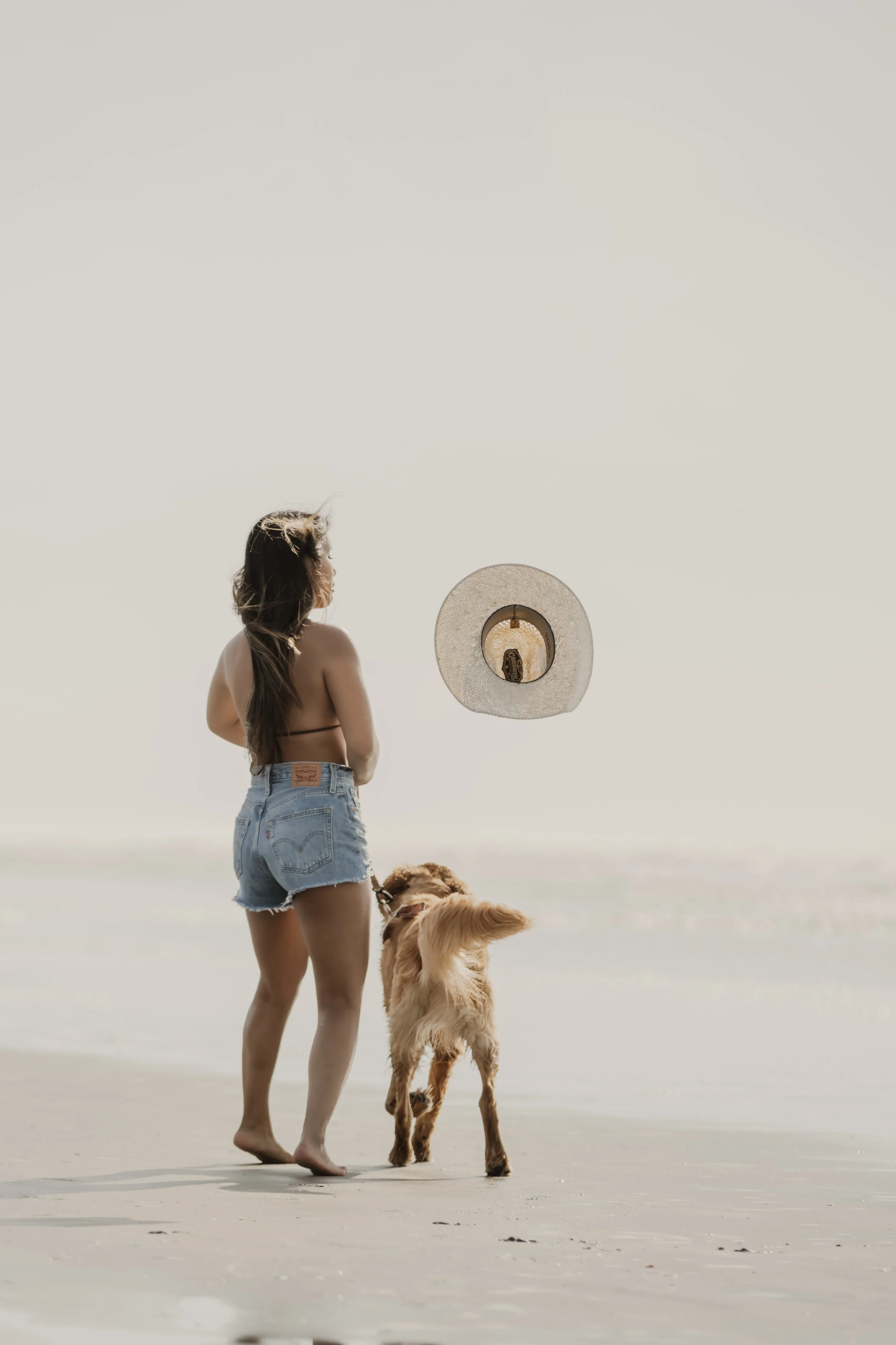 A woman in a bikini top and denim shorts walking on the beach with her dog during daytime, with a hat hanging in the air.