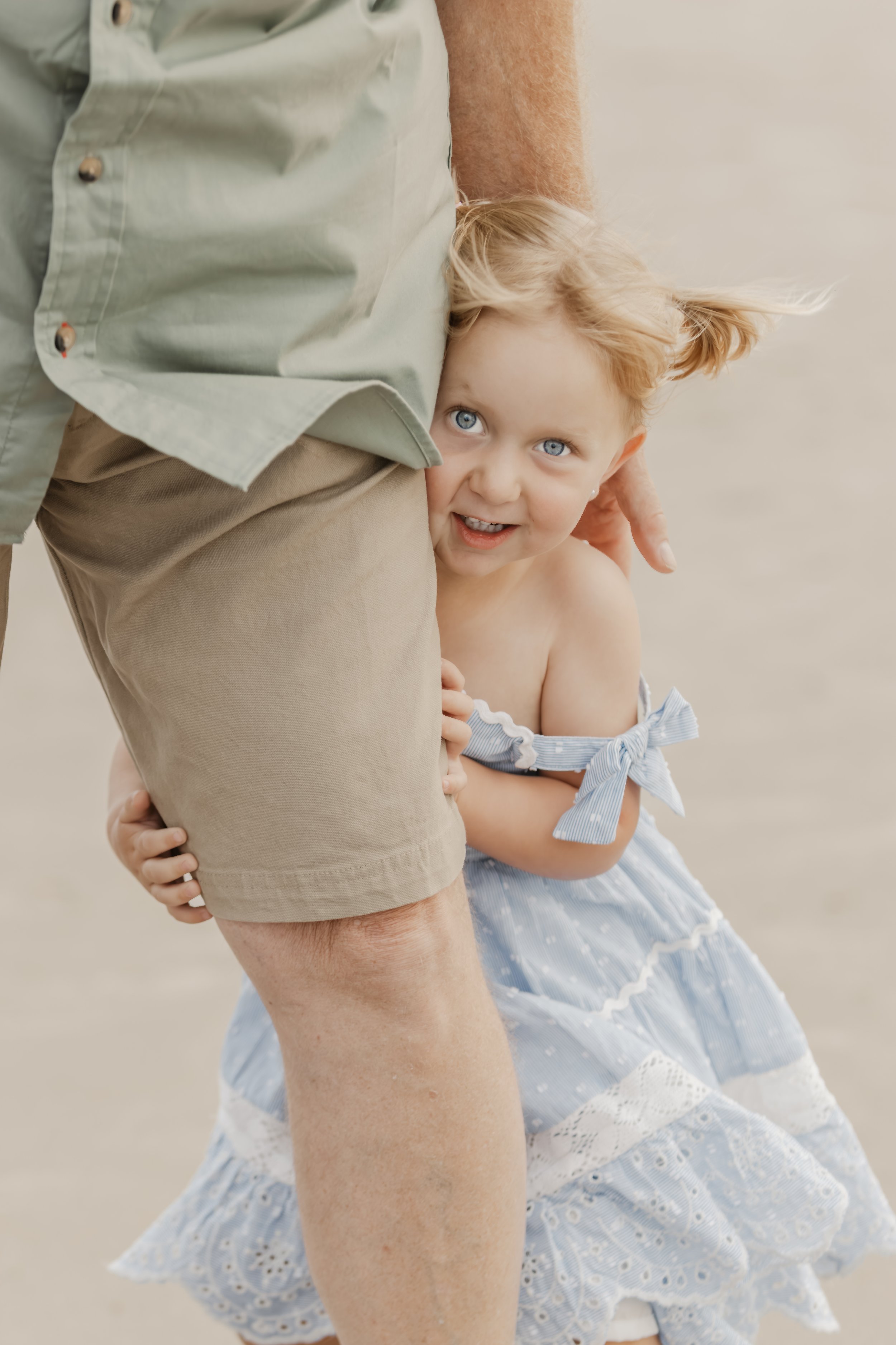 A young girl with blue eyes and blonde hair holding onto an adult's leg, partially hidden behind a knee-length khaki shorts worn by the adult, on a sandy beach.