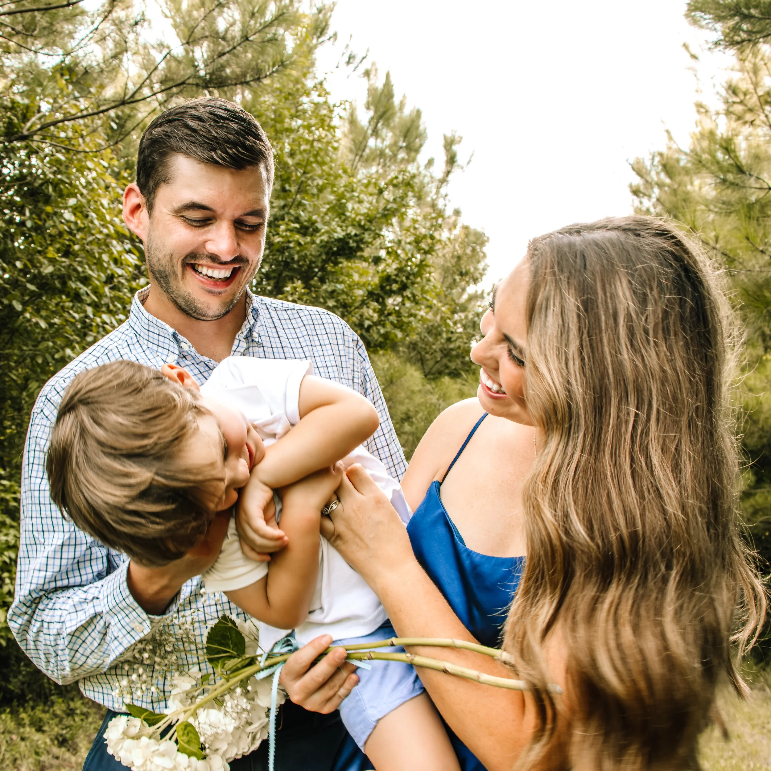 A happy family of three, a father, mother, and young daughter, outdoor in a park with trees. The father is holding the daughter, who is smiling and playing with the mother, who is holding flowers.