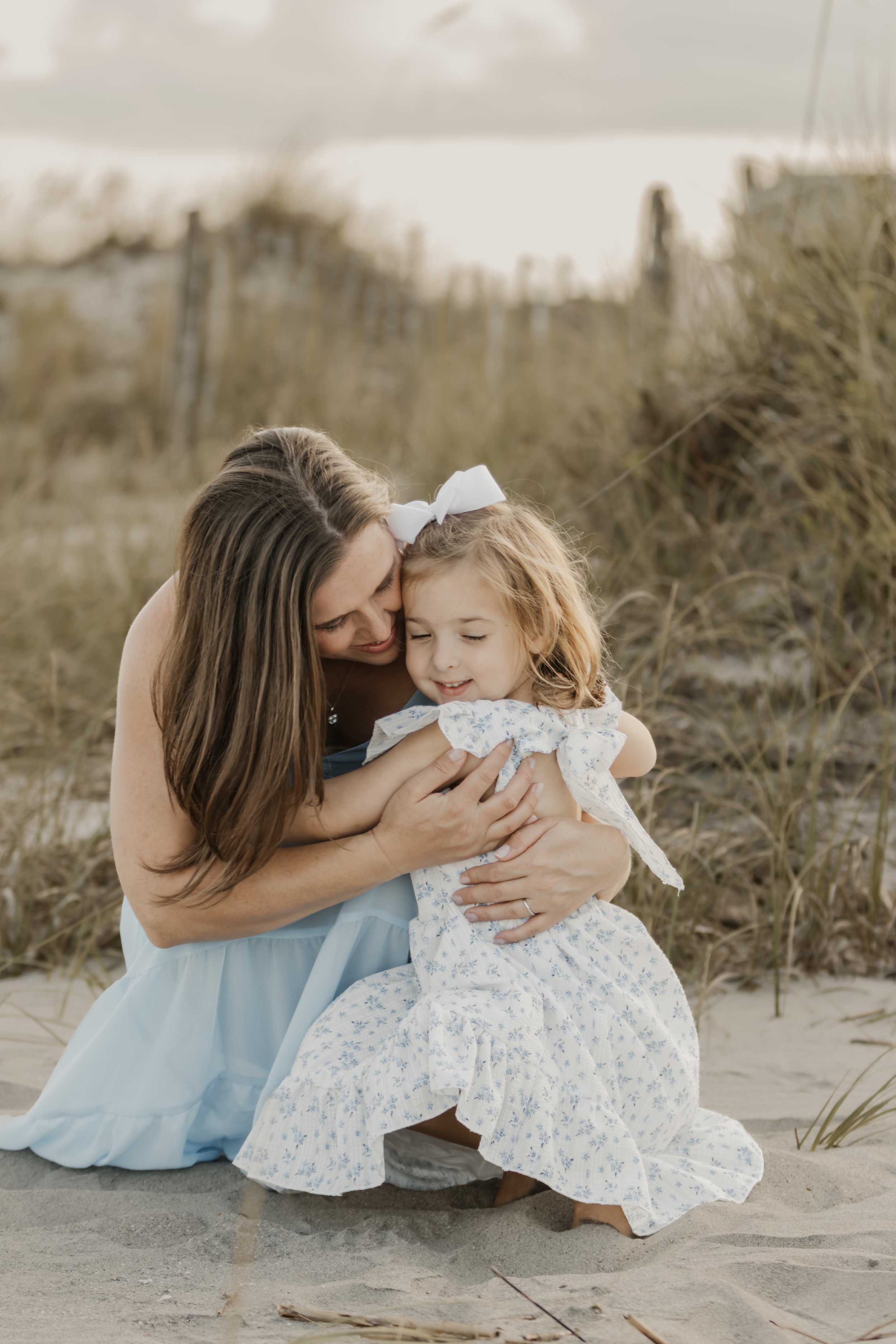 A woman and a girl hugging on a sandy beach with dunes and tall grass in the background.
