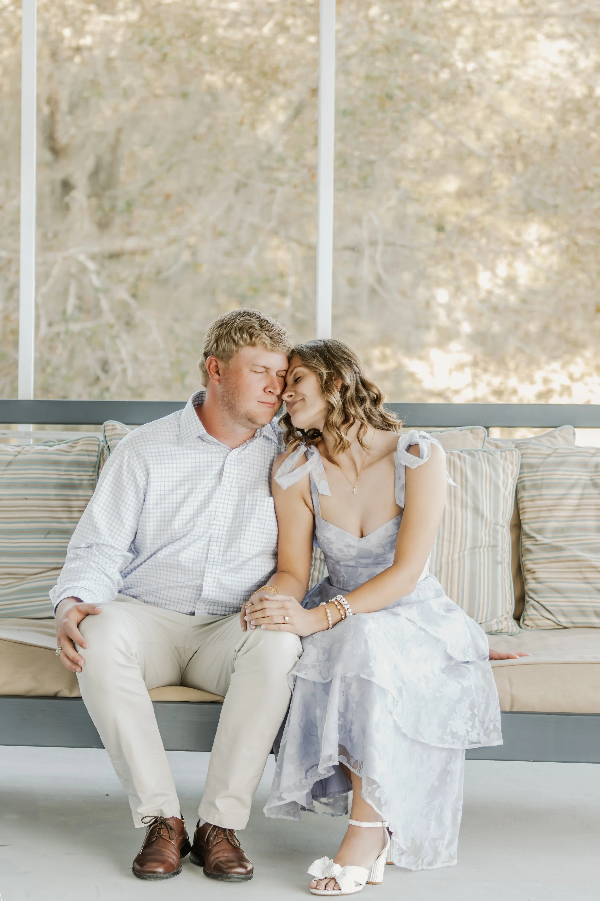 A young couple sitting close together on a cushioned bench with striped pillows, sharing an intimate moment with their foreheads touching, in front of large window with a blurred outdoor scene.