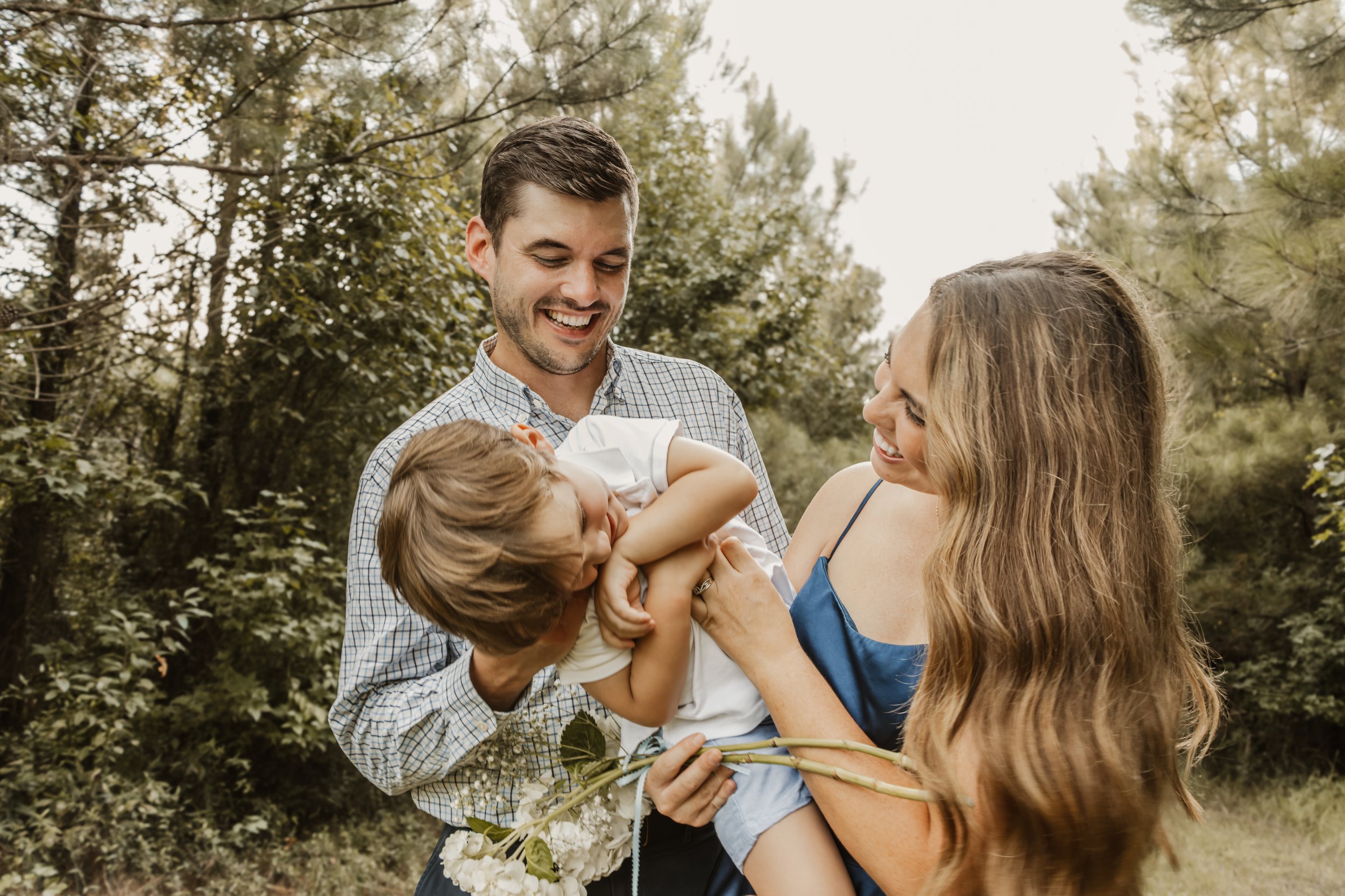 A family of three, a father, mother, and their young son, are outdoors in a wooded area. The father and mother are smiling as the son laughs and plays with them. The mother is holding a bouquet of white flowers.