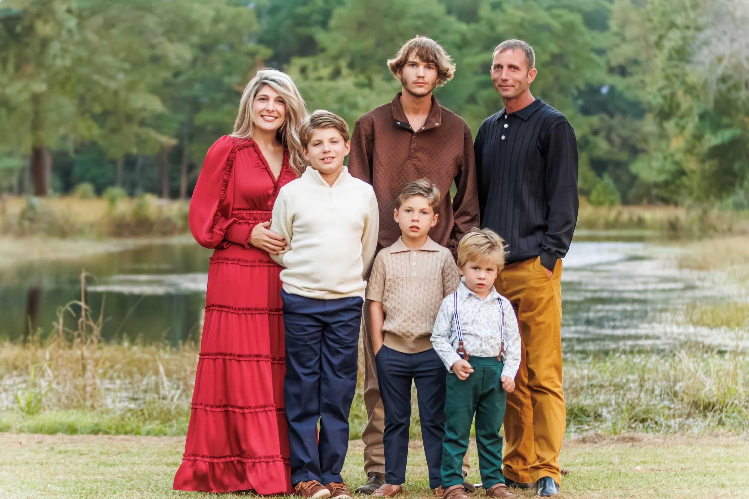 Family of six standing outdoors near a lake with trees in the background. The woman wears a red dress, the men and boys wear casual shirts and pants, and the children have varying hairstyles and expressions.