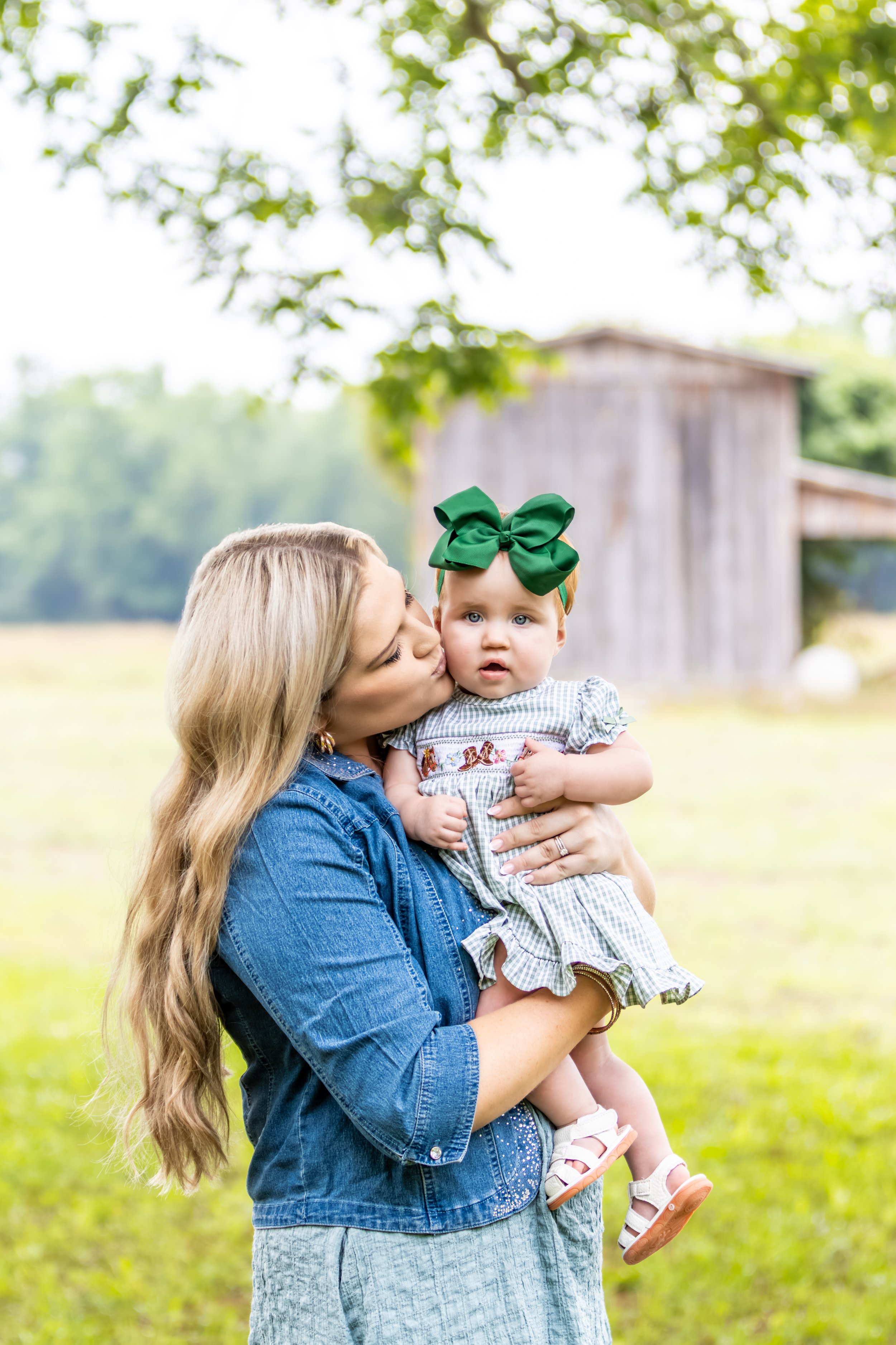 A woman with long blonde hair in a denim jacket holds a young girl with a large green bow in her hair, outdoors with a wooden barn and trees in the background.