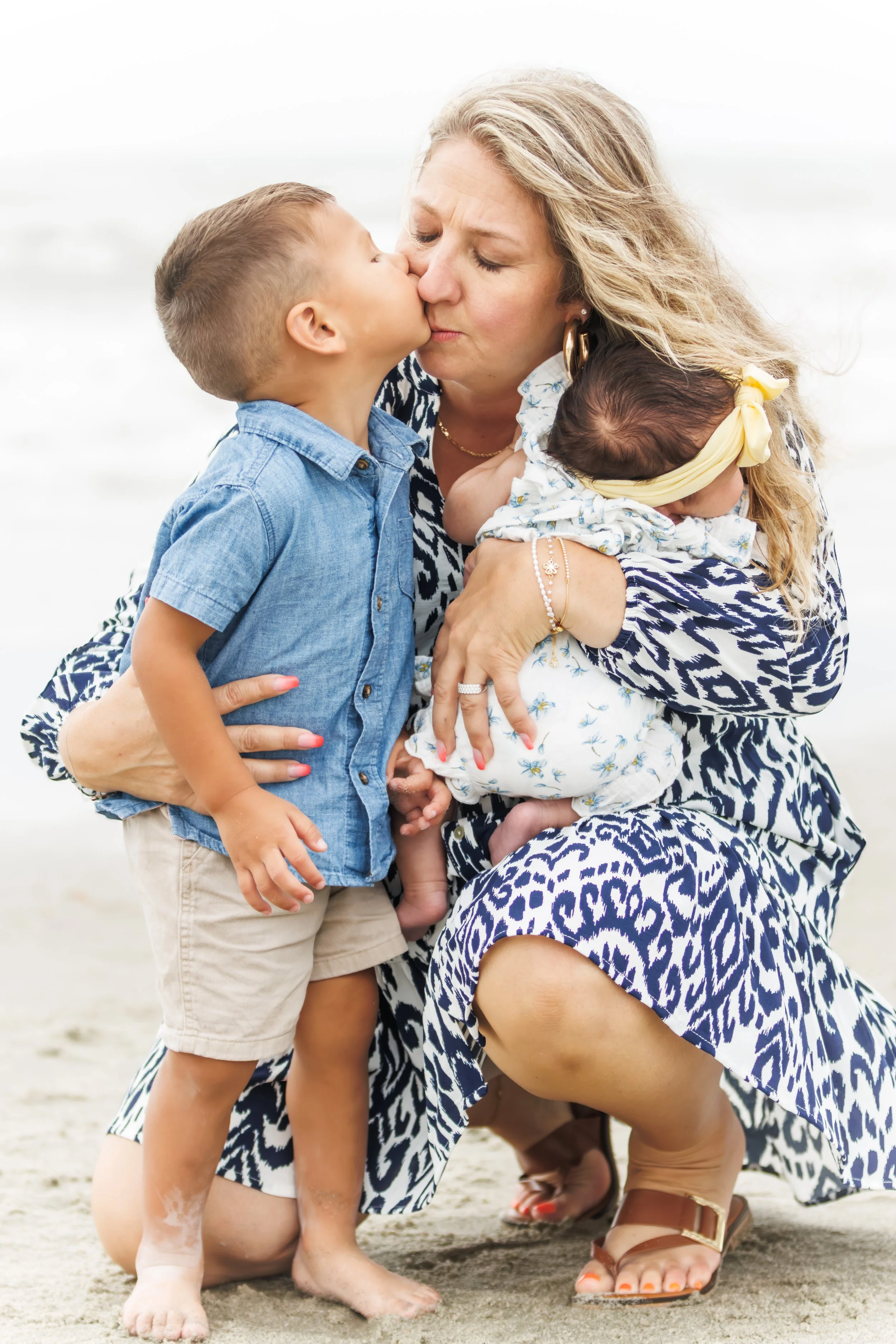 A woman on a beach kissing a young boy, with a toddler girl in her arms, all three dressed in casual summer clothing.