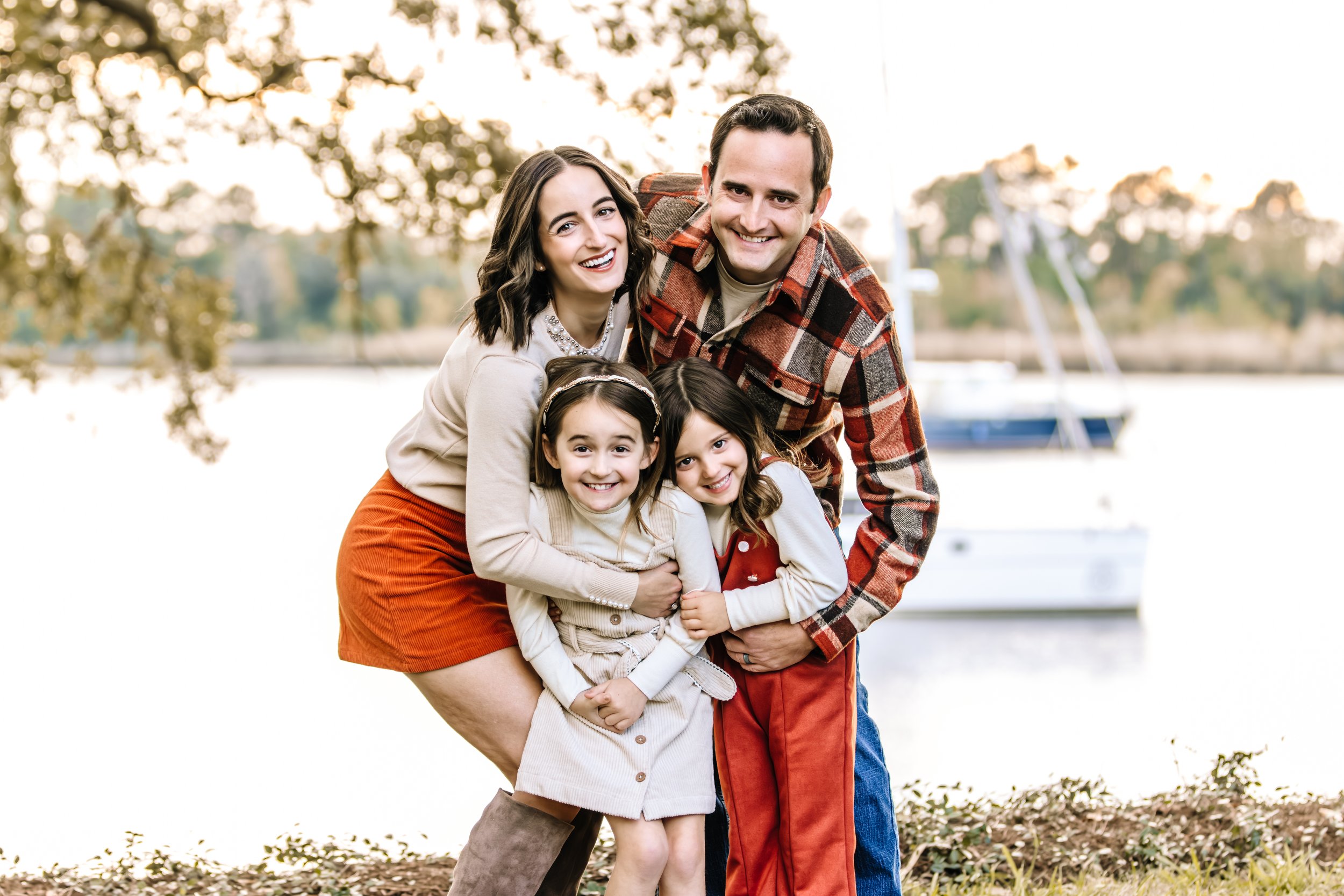 A happy family of four, with two young girls, posing outdoors near a body of water with boats in the background, during sunset.
