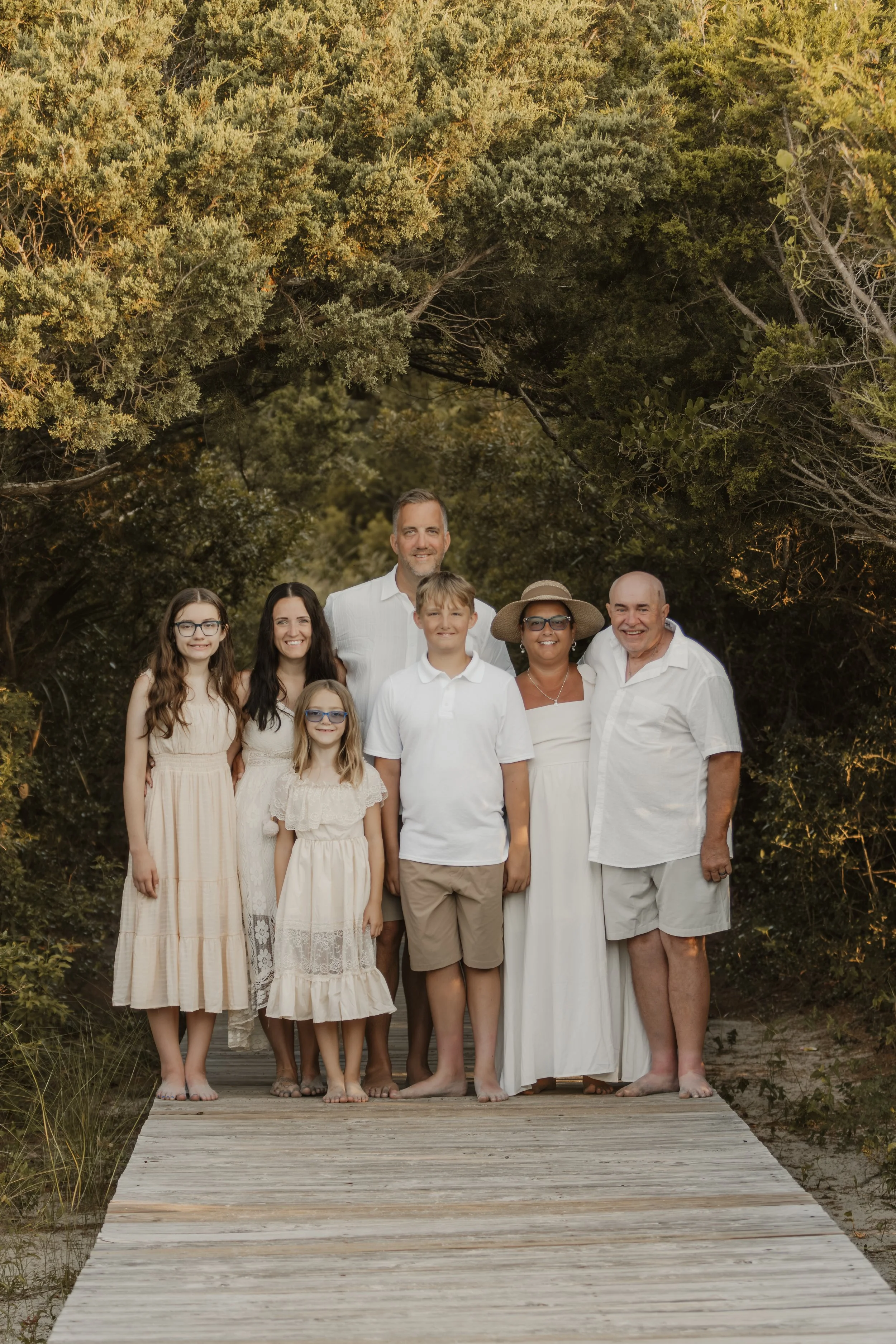 A family of eight standing on a wooden pathway outdoors, surrounded by greenery and trees, dressed in white and neutral clothing.