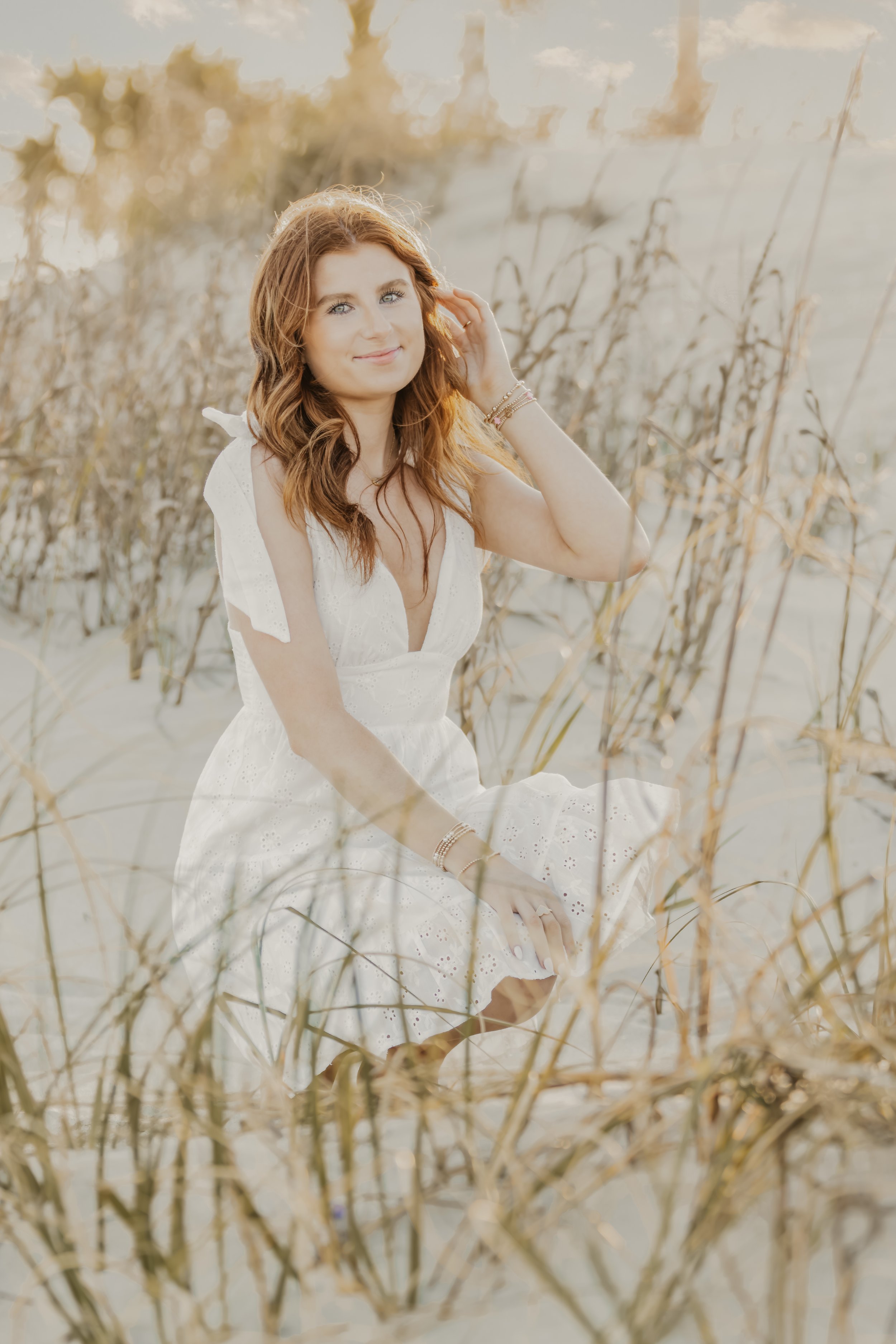 A woman with red hair, wearing a white dress, sitting on sand among tall dry grass, smiling and touching her hair in a natural outdoor setting at sunset.