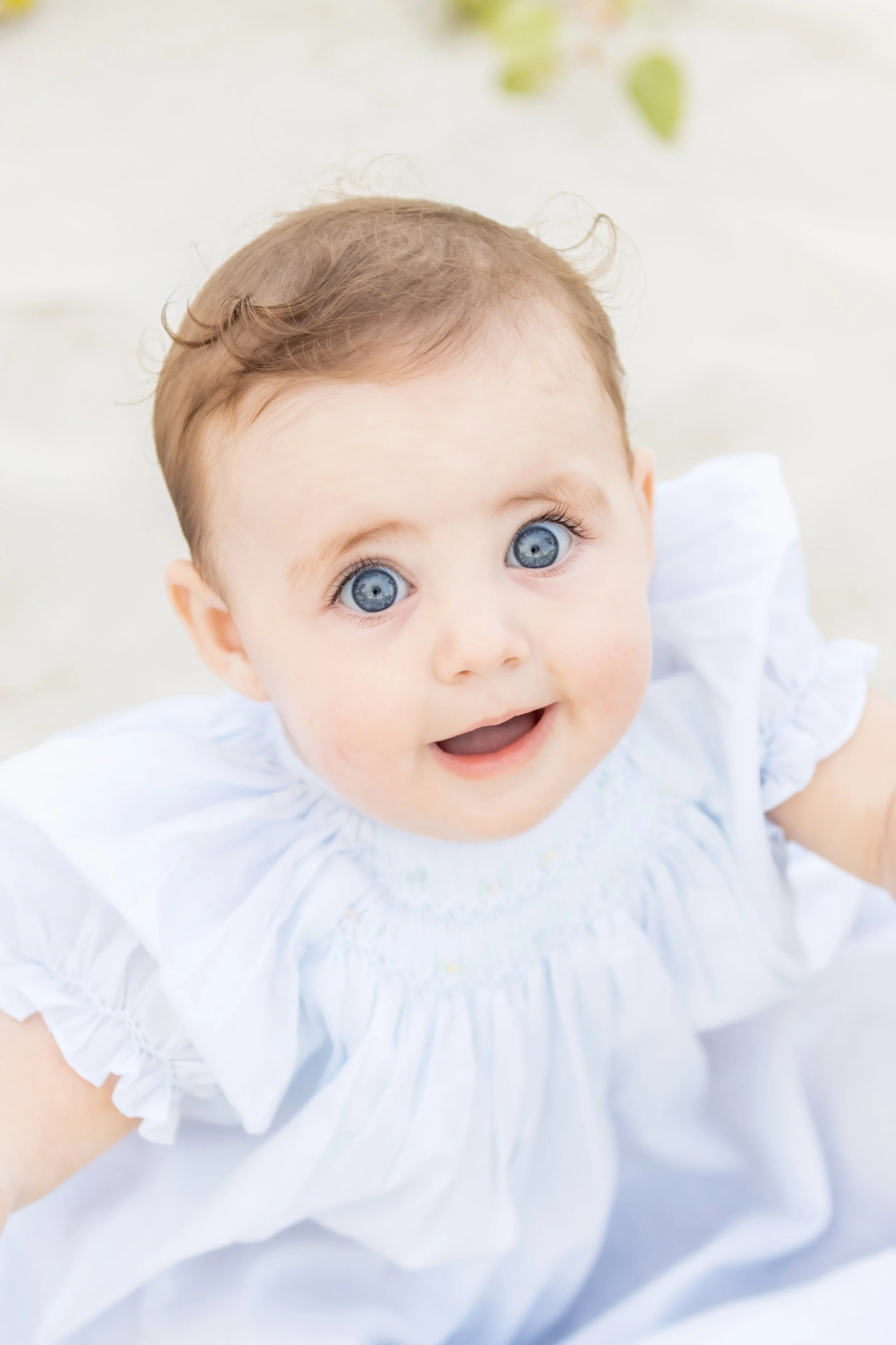 A baby with blue eyes and light brown hair, wearing a white dress, looking up with an open-mouthed smile.