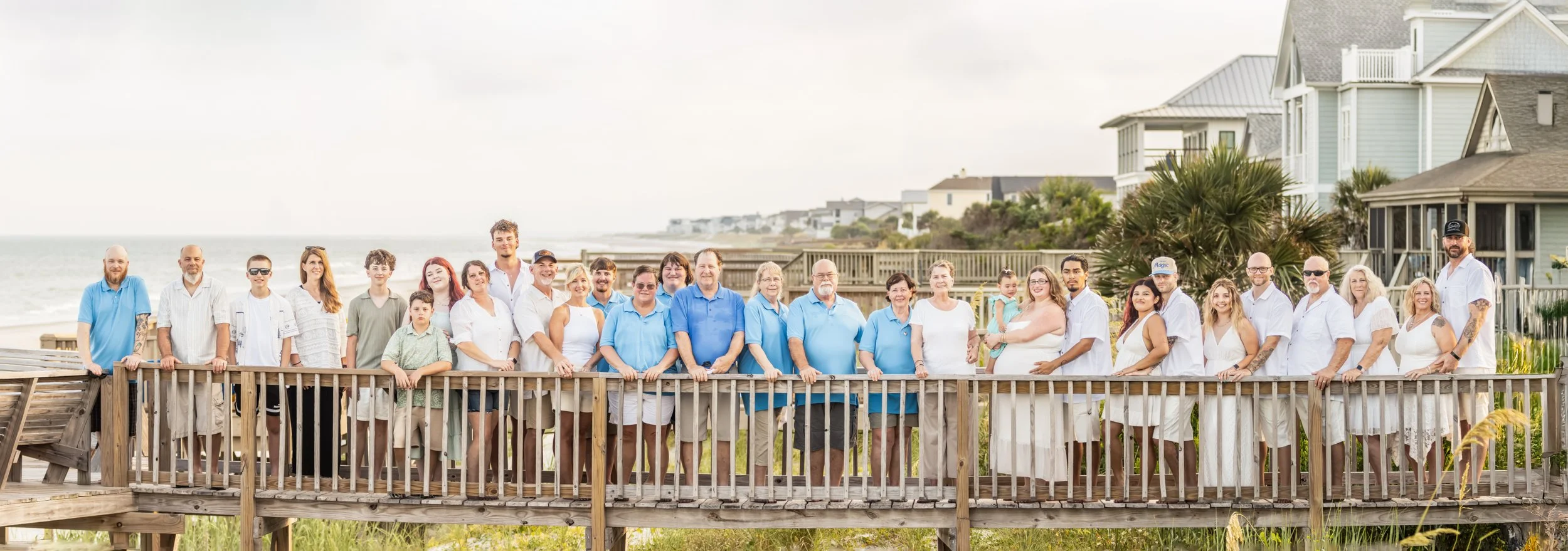 Group of people standing on a wooden deck near the beach, with houses and the ocean in the background.