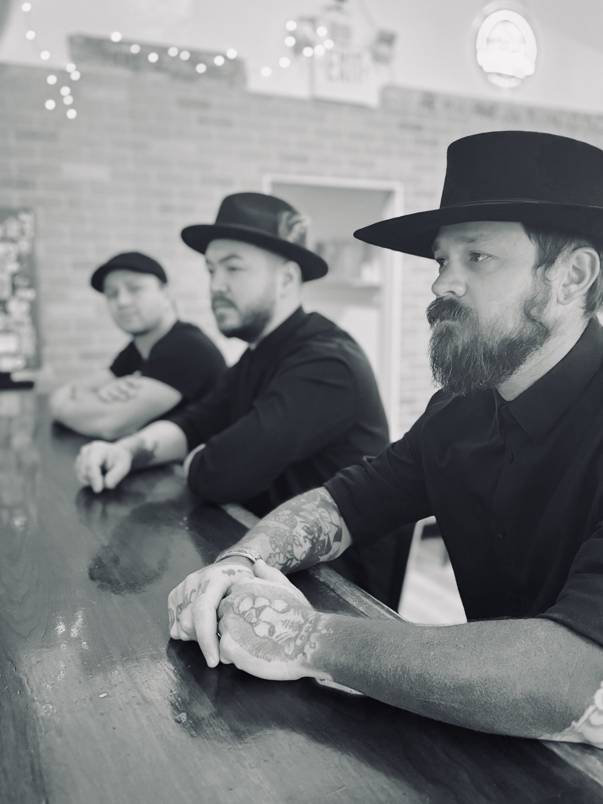 Three men with tattoos wearing black shirts and hats sitting at a bar or counter in a casual setting with string lights and brick wall in the background.
