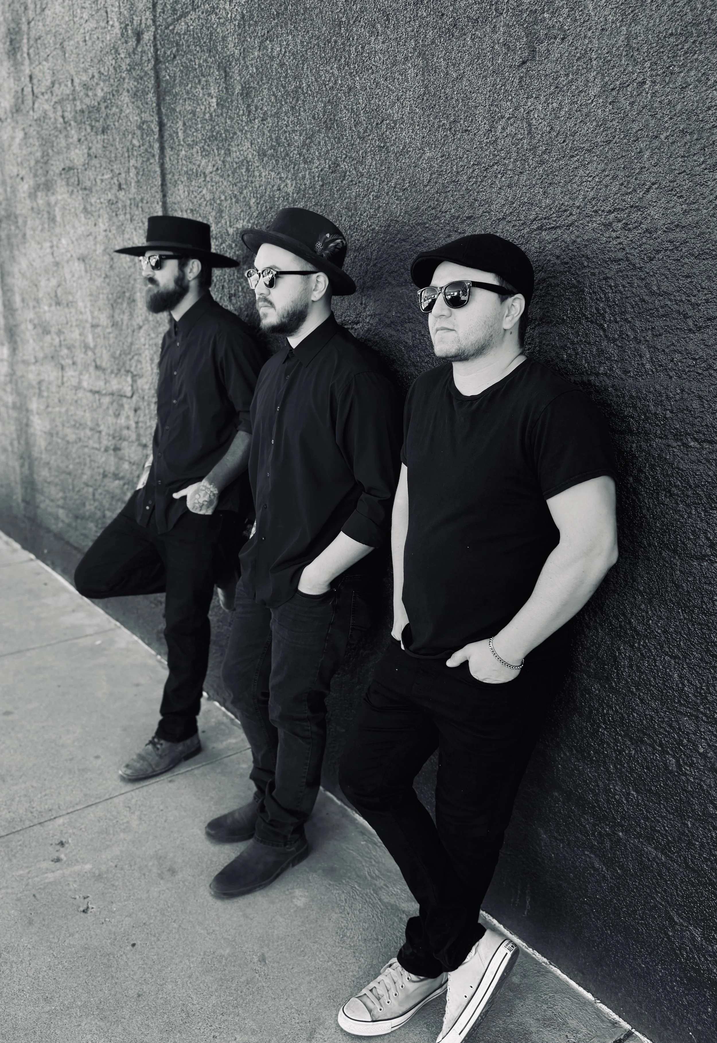 Three men standing against a textured wall, wearing dark clothing and sunglasses, with a casual pose.