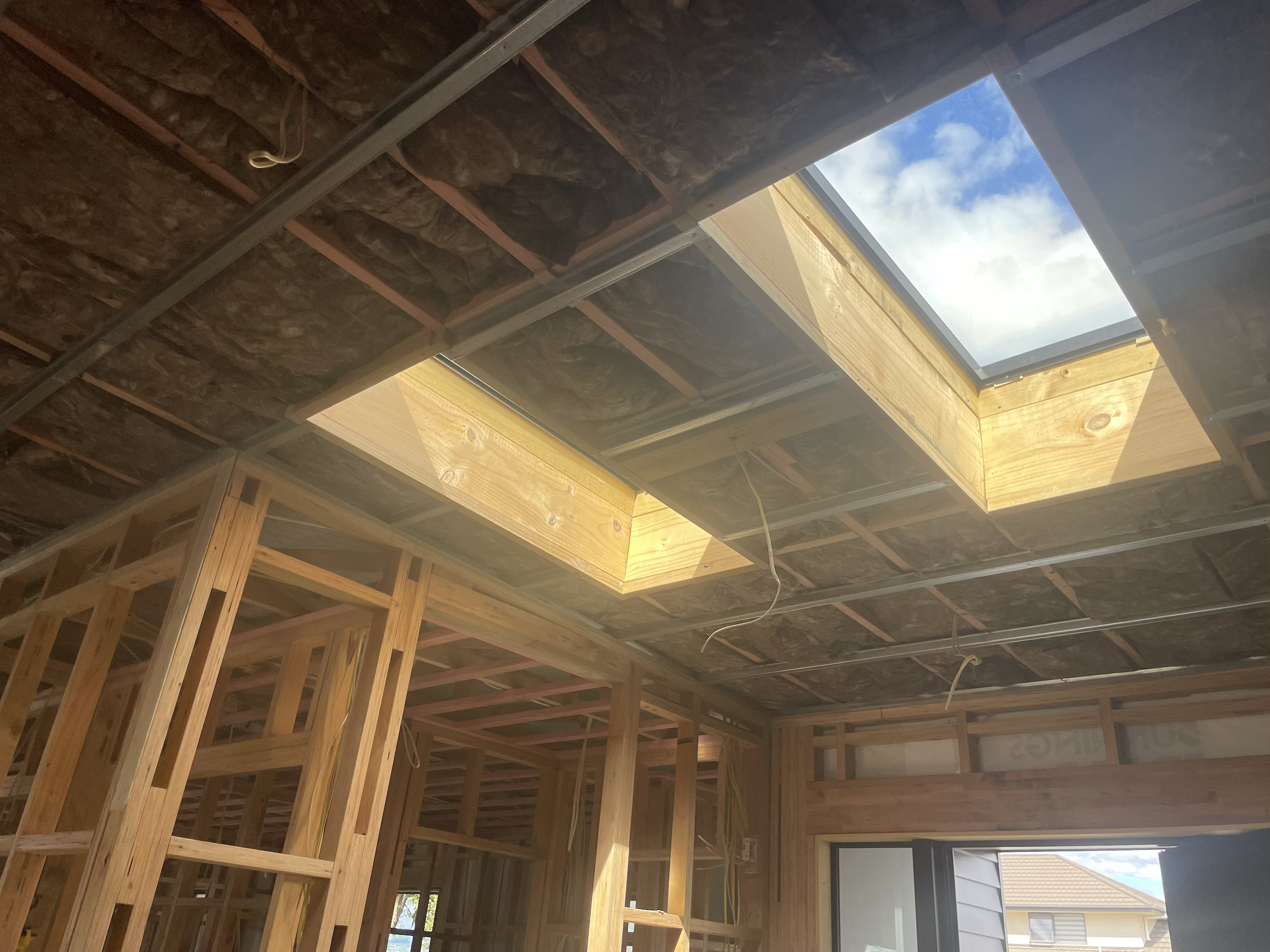 Interior of a house under construction with exposed wood framing and two skylights in the ceiling, showing a partly cloudy sky outside.