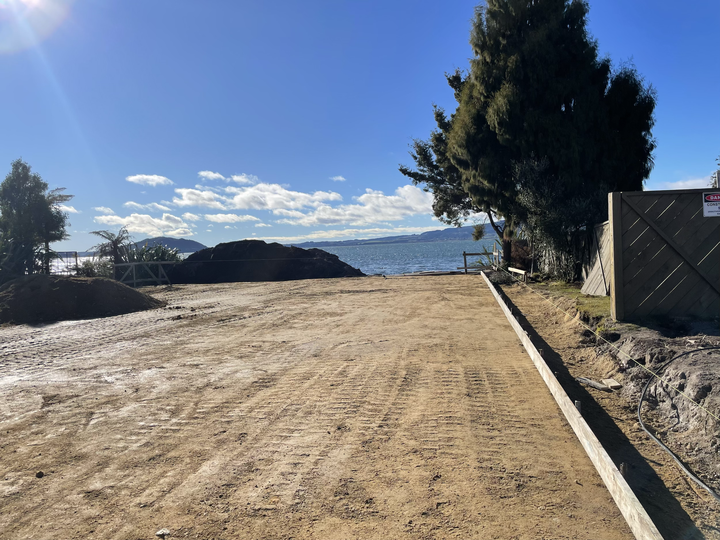 Unpaved construction area near a body of water with a large tree and a fence on the right side, and a hillside in the background under a partly cloudy sky.