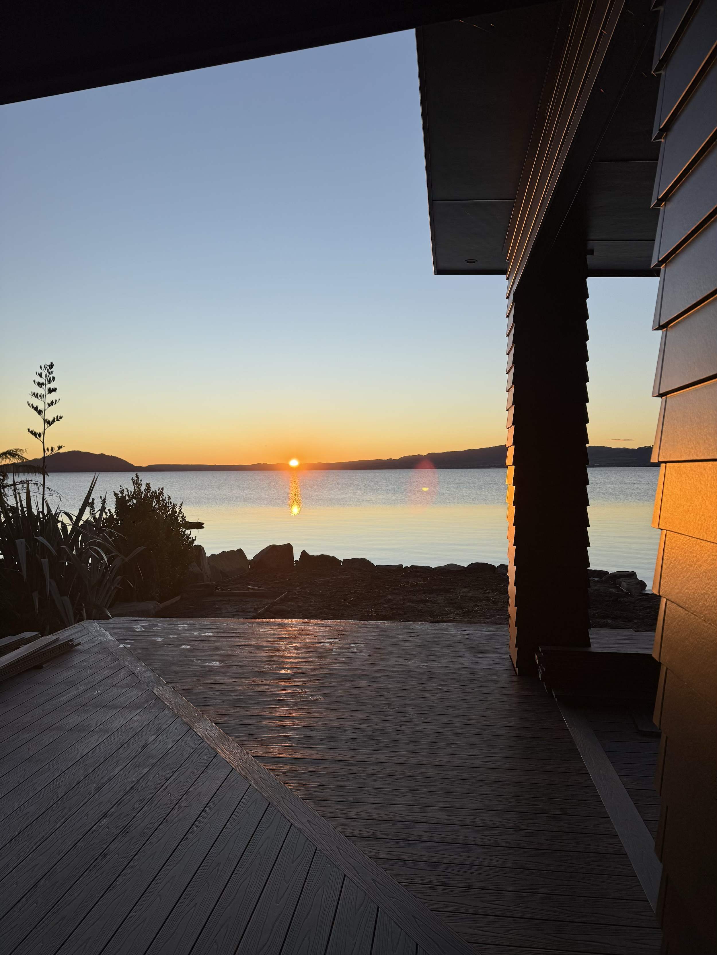 Sunset over a calm body of water viewed from a wooden deck, with part of a modern building's patio in the foreground.