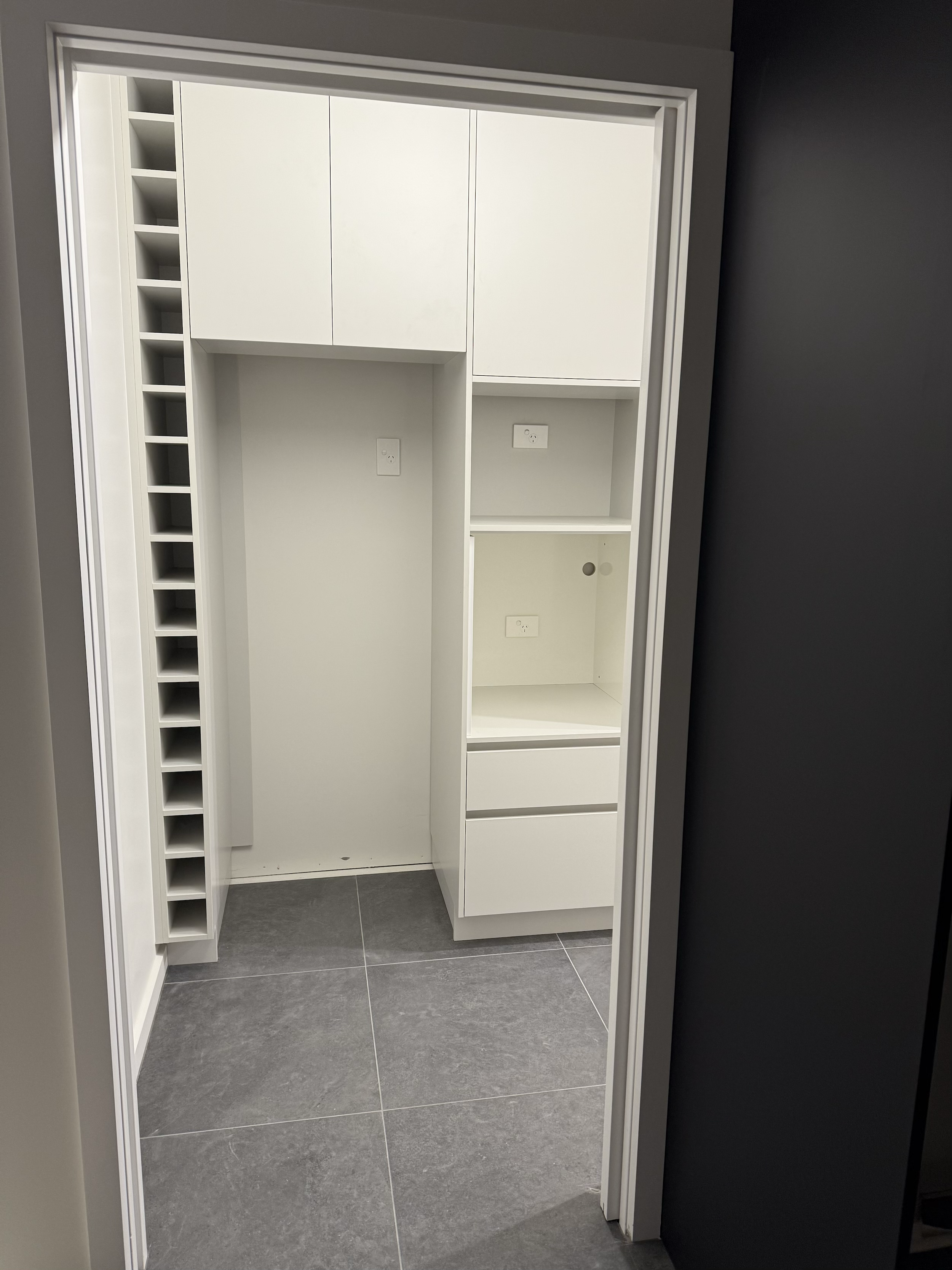 Empty white walk-in pantry with multiple cabinets and drawers, and a wine rack on the left side, with gray tiled flooring.
