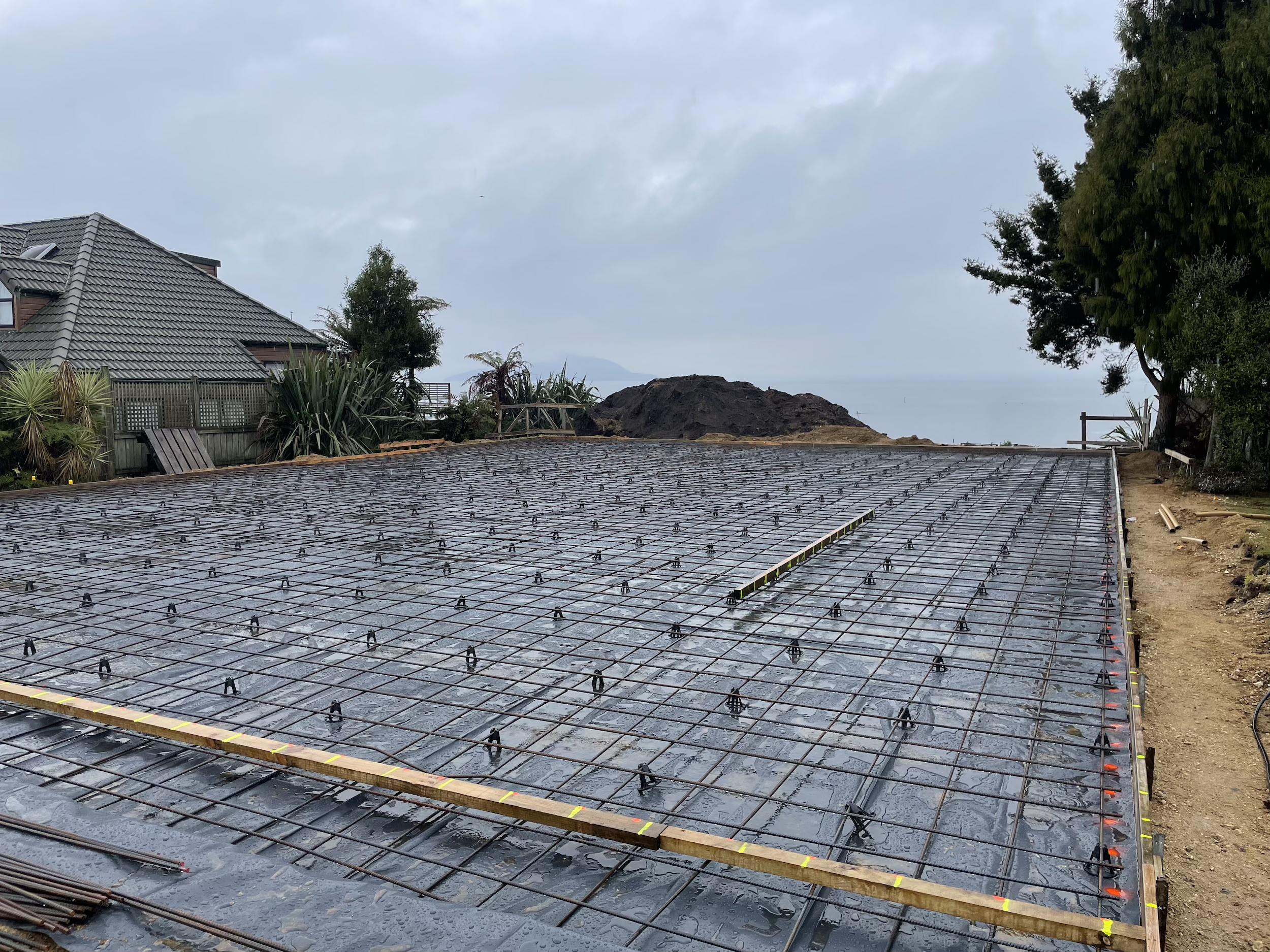 Construction site with a steel reinforcement grid laid out for a concrete slab, overlooking the ocean on a cloudy day.