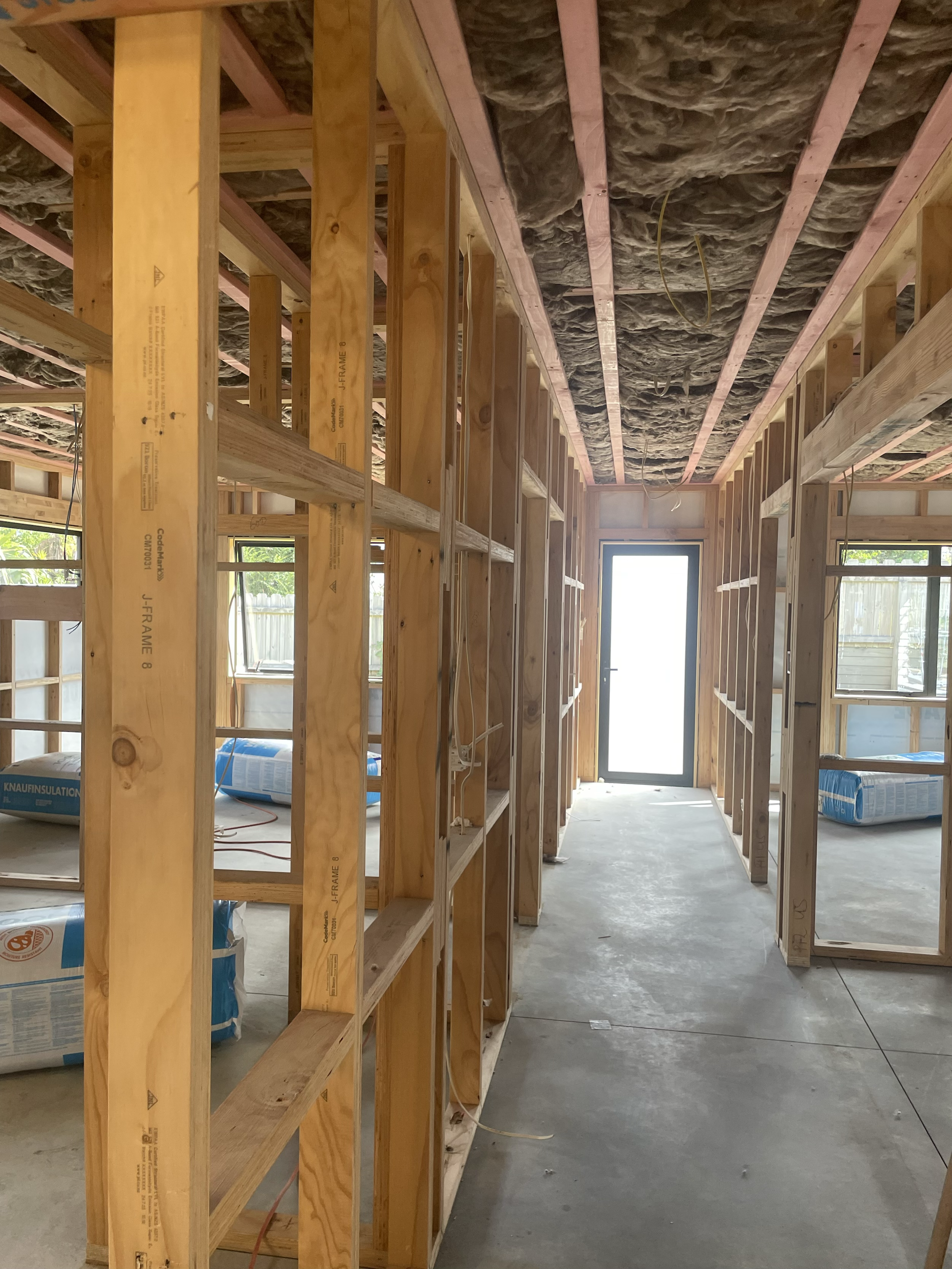 Interior of a house under construction with wooden framing, insulation on the ceiling, and a glass door at the end of the hallway.